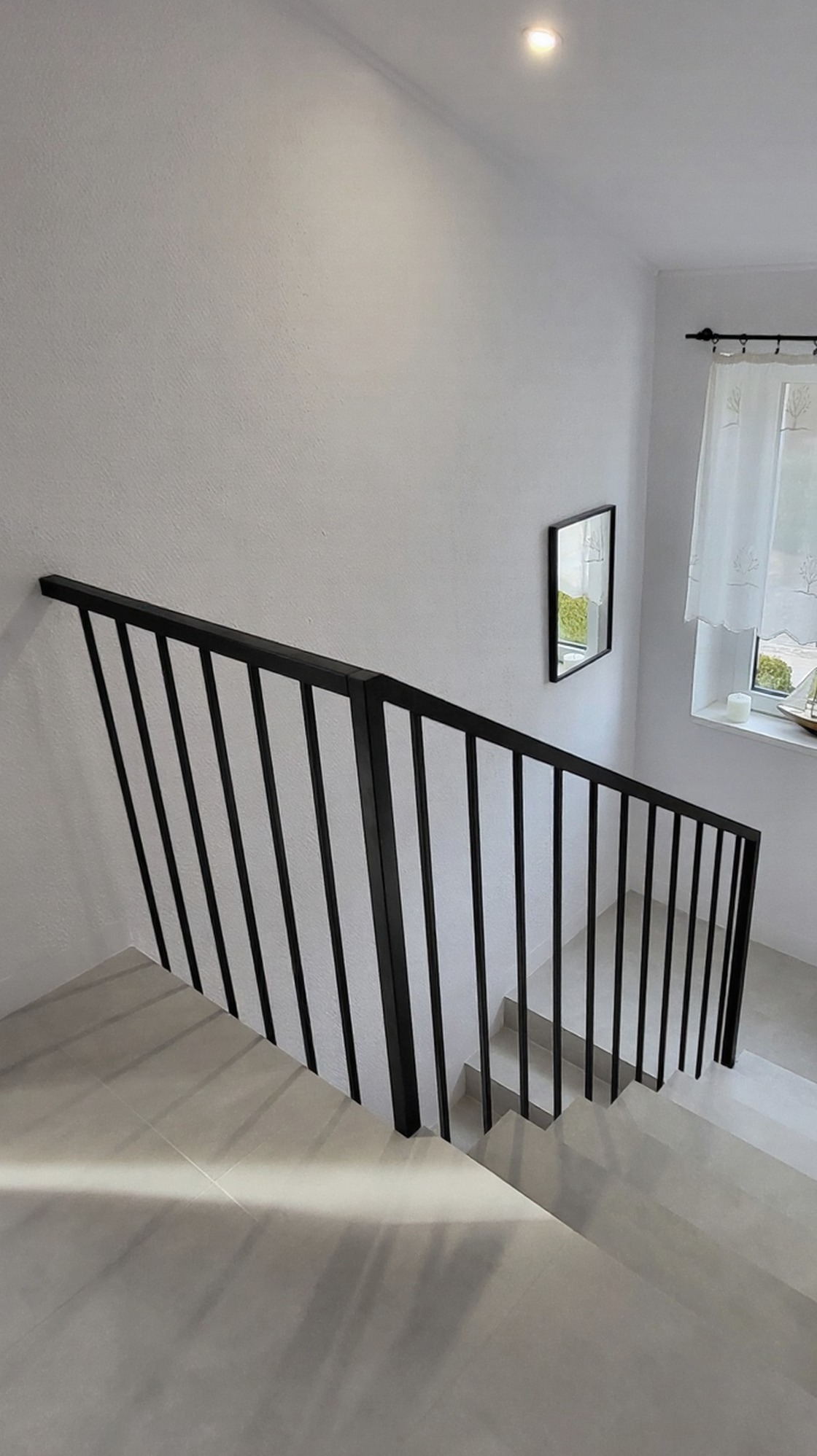 Minimalistyczna, czarna balustrada schodowa z pionowymi prętami, zamocowana do ściany. Widok z góry na jasne schody, okno z firanką i lustro w tle. Nowoczesny design.