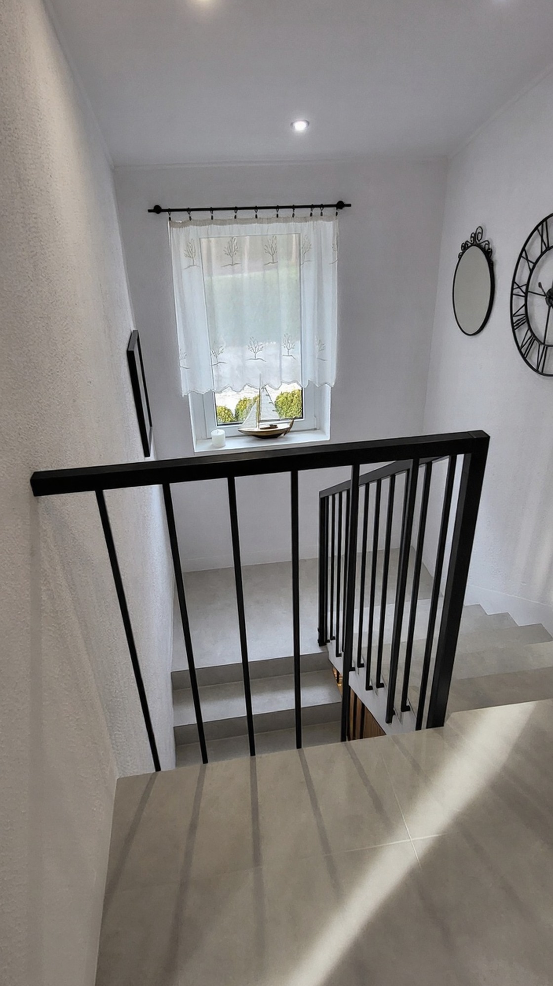 Minimalistyczne schody z czarną, metalową balustradą z pionowymi prętami. Widok z góry na jasne stopnie i balustradę, okno z dekoracją w tle.