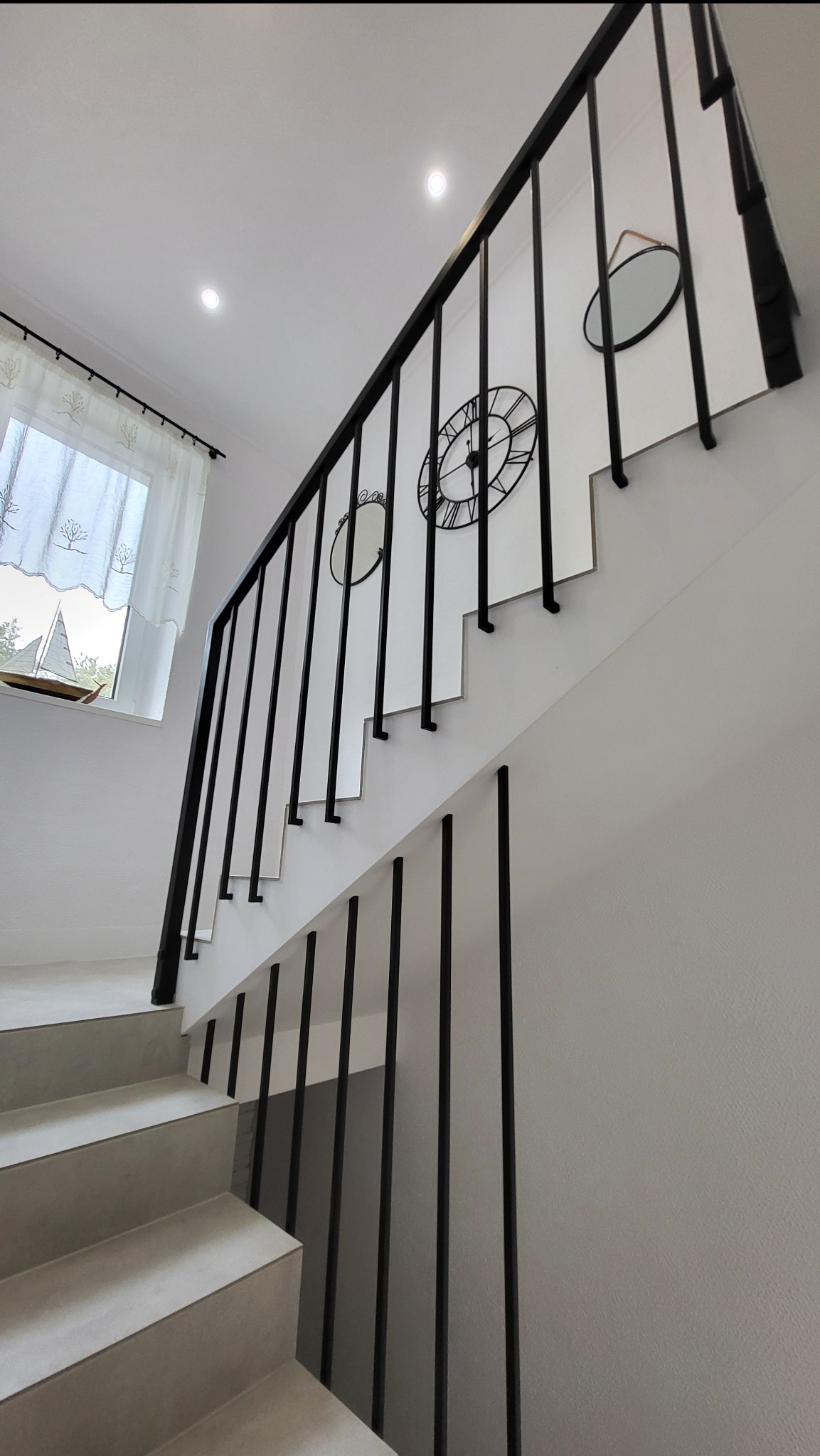 Nowoczesne schody z czarną, minimalistyczną balustradą z pionowych prętów, zamocowaną do ściany. Na ścianie dekoracje: zegar i lustra. W tle okno z firanką.
