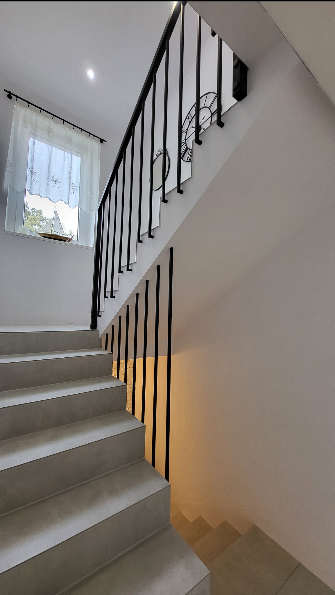 Minimalistyczne schody z czarną, metalową balustradą z pionowymi tralkami, mocowanymi do ściany. W tle okno z dekoracyjną firanką i zegar na ścianie.