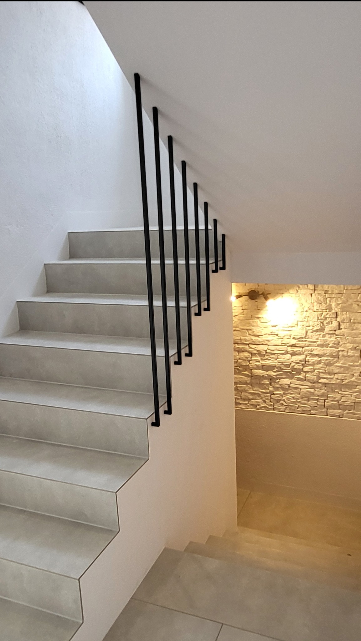 Minimalistyczne schody z czarną, metalową poręczą mocowaną do ściany. Jasne stopnie i ściany, oświetlona ściana z kamienia w tle. Nowoczesny design.