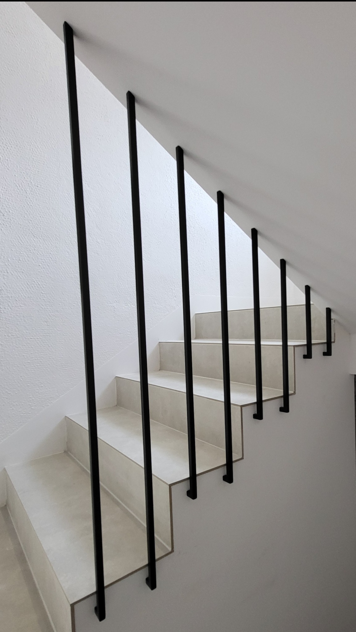 Minimalistyczna balustrada schodowa z czarnymi, pionowymi tralkami mocowanymi do ściany, bez dolnego profilu, na tle jasnych schodów i białych ścian.