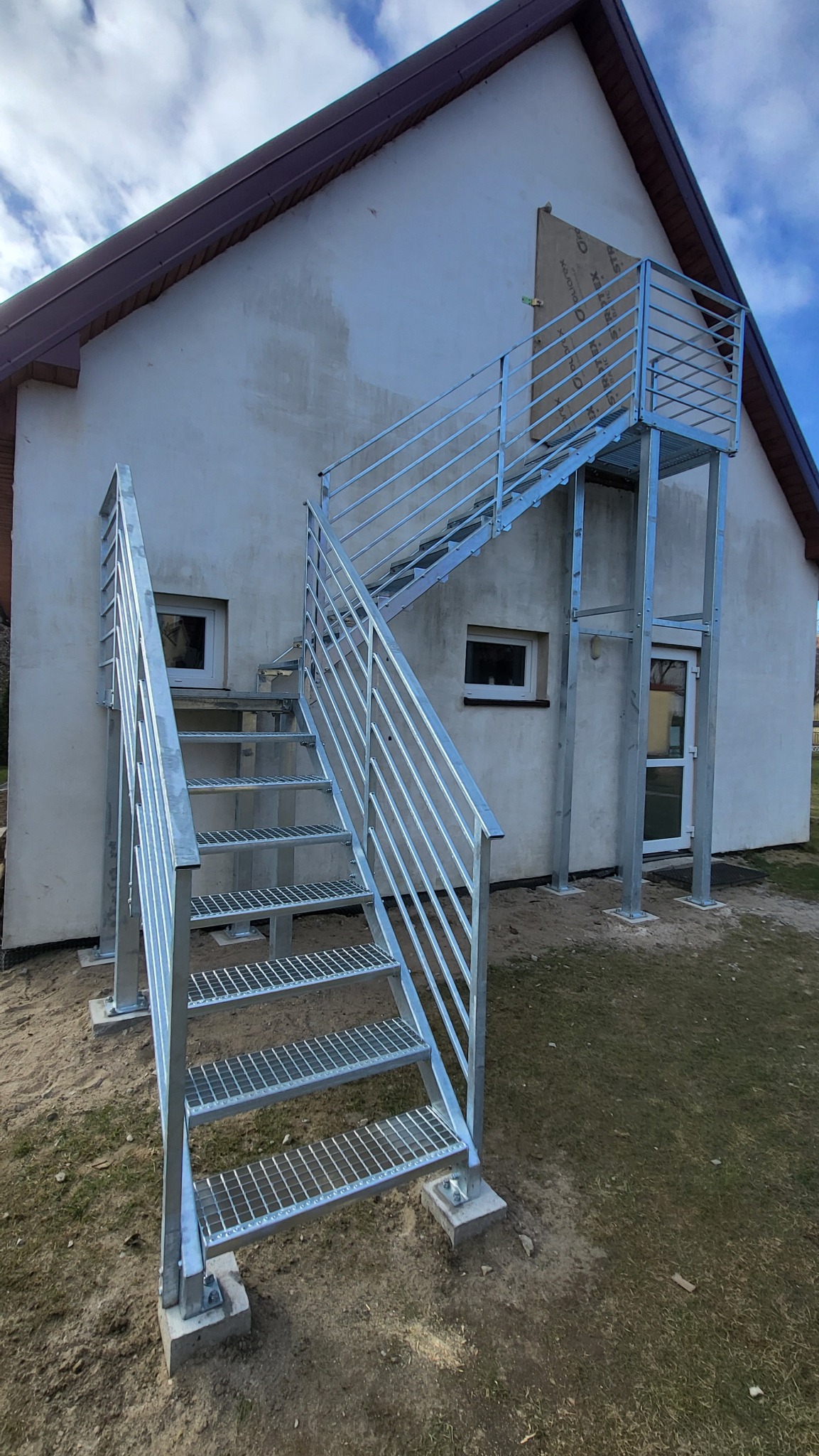 Zewnętrzne schody metalowe z balustradą, ocynkowane ogniowo, prowadzące do drzwi na piętrze budynku. Stopnie z kraty WEMA, filary i słupki z metalowych profili.