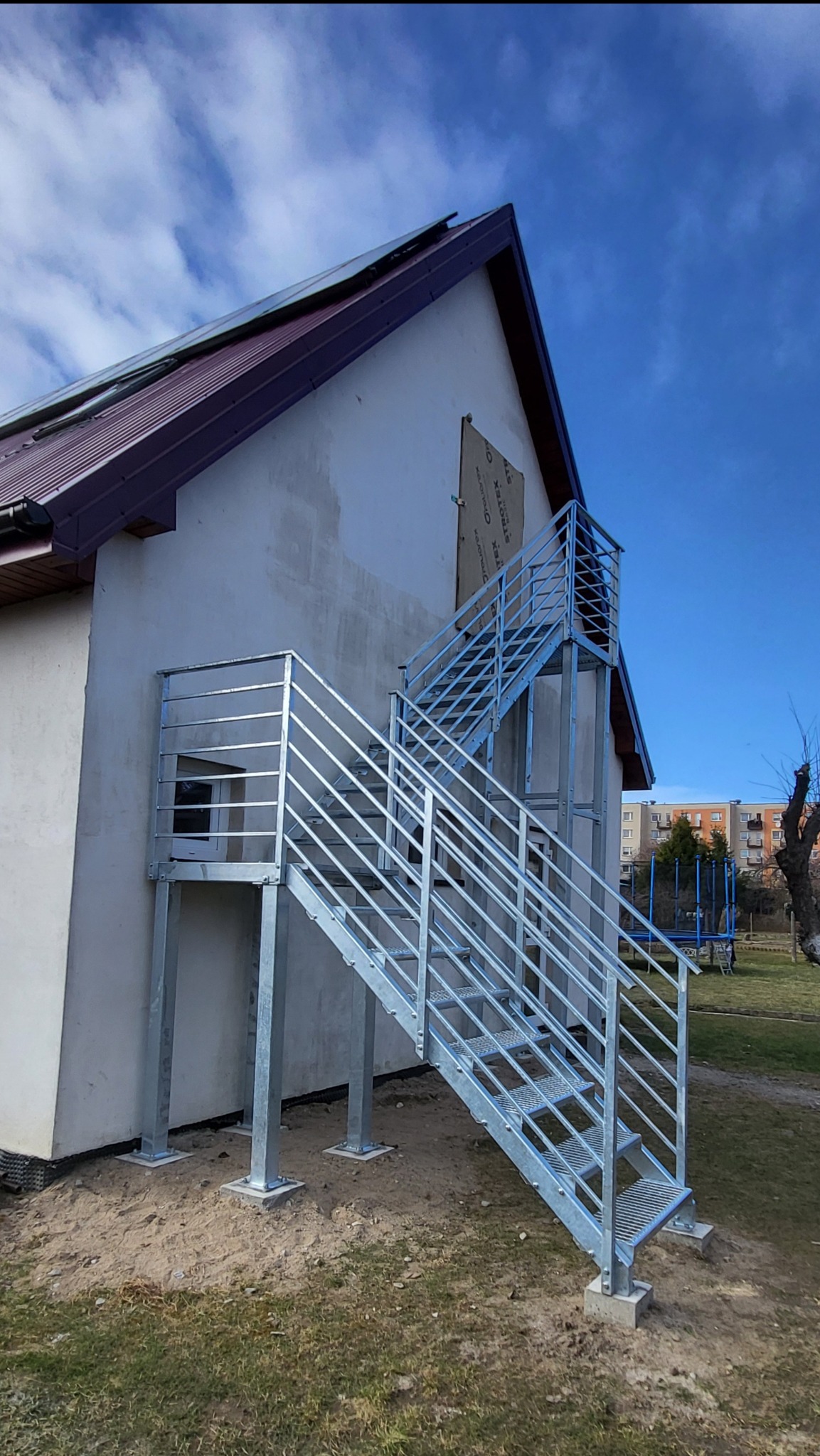 Zewnętrzne schody metalowe z balustradą, ocynkowane ogniowo, prowadzące do wejścia na piętro budynku. Stopnie z kraty WEMA, słupki i wypełnienie balustrady z profili.