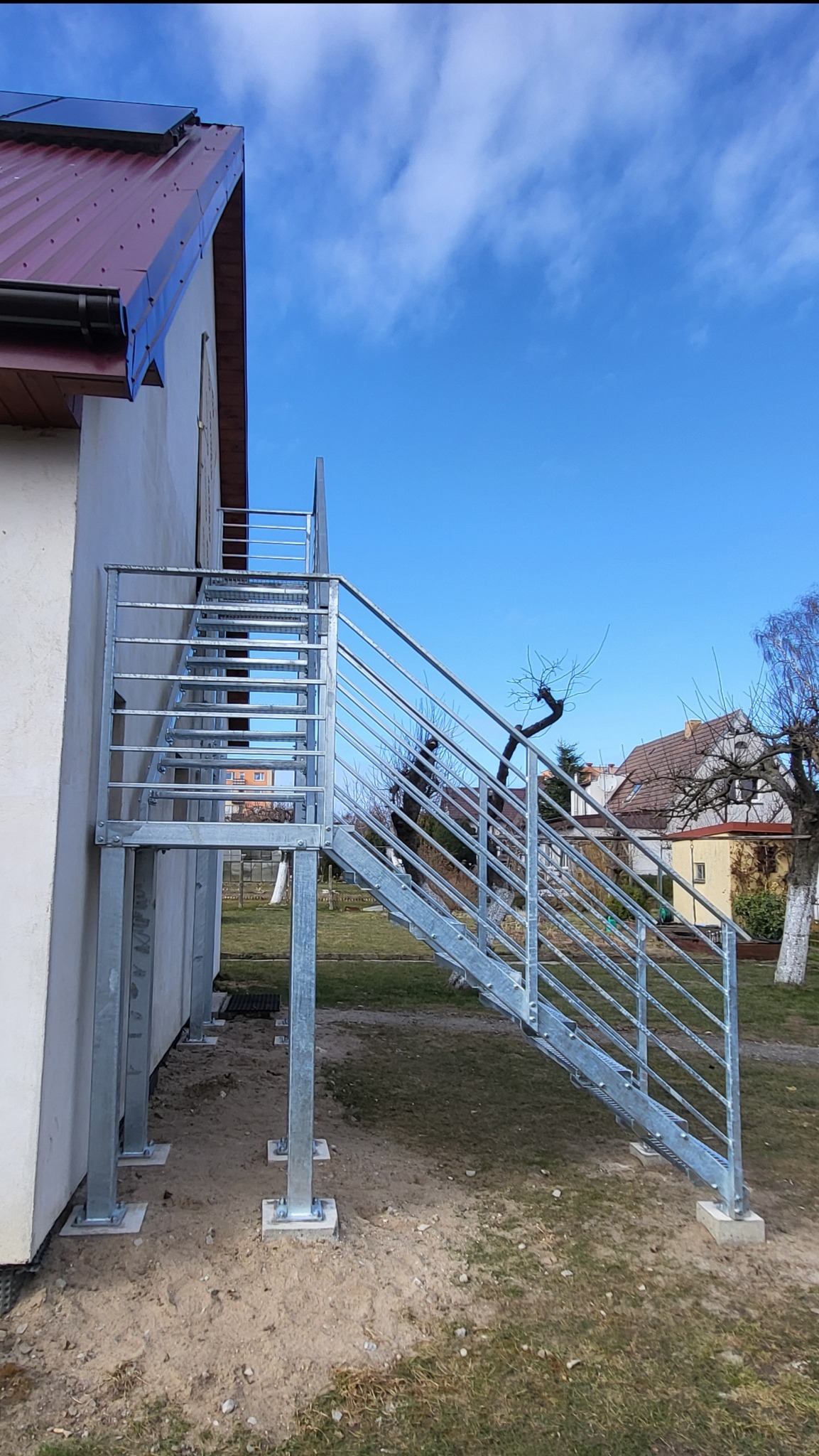 Zewnętrzne schody metalowe z balustradą, ocynkowane ogniowo, prowadzące do budynku. Stopnie z kraty WEMA, słupki i wypełnienie balustrady z profili stalowych.
