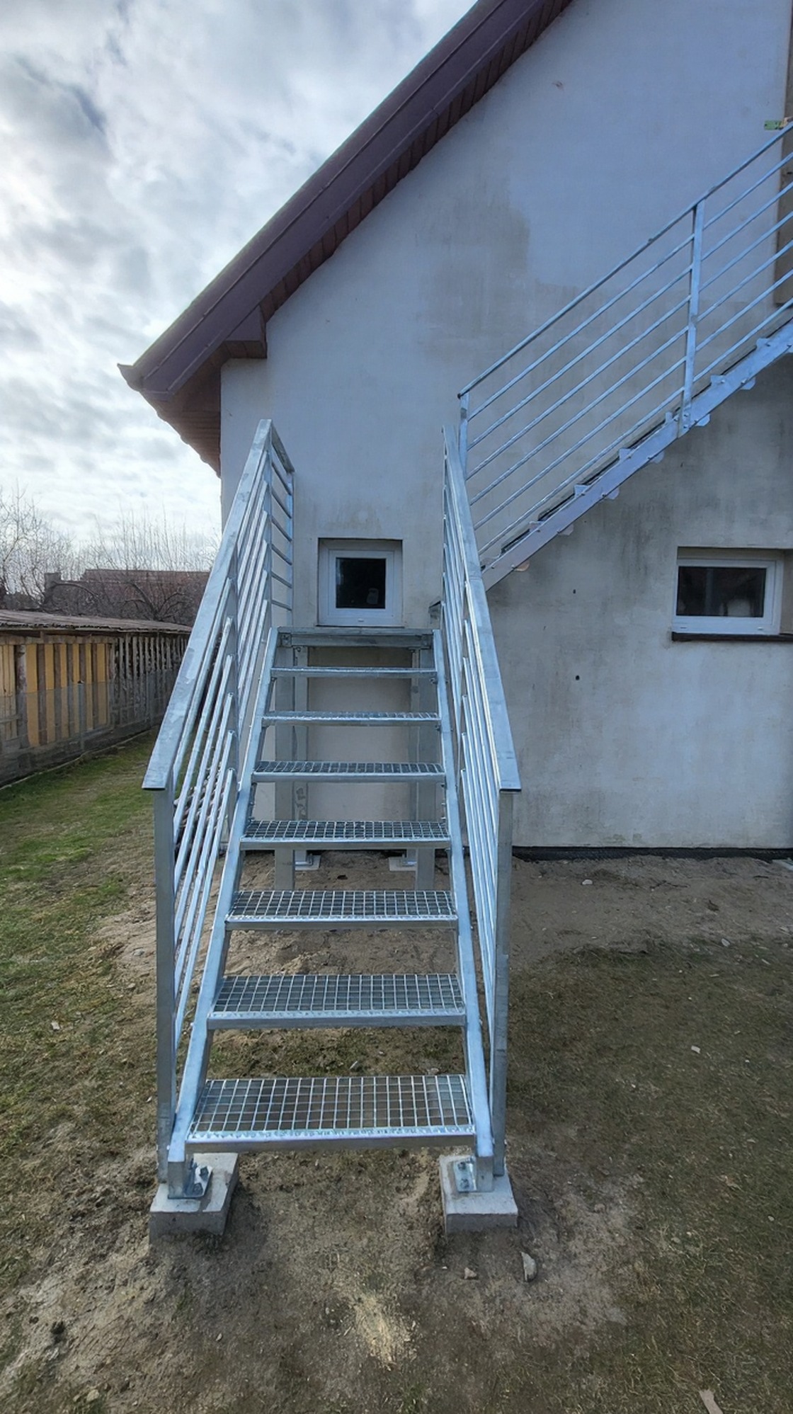Zewnętrzne schody metalowe z balustradą, ocynkowane ogniowo, prowadzące do wejścia na piętro budynku. Stopnie z kraty WEMA, filary i słupki balustrady z profili stalowych.