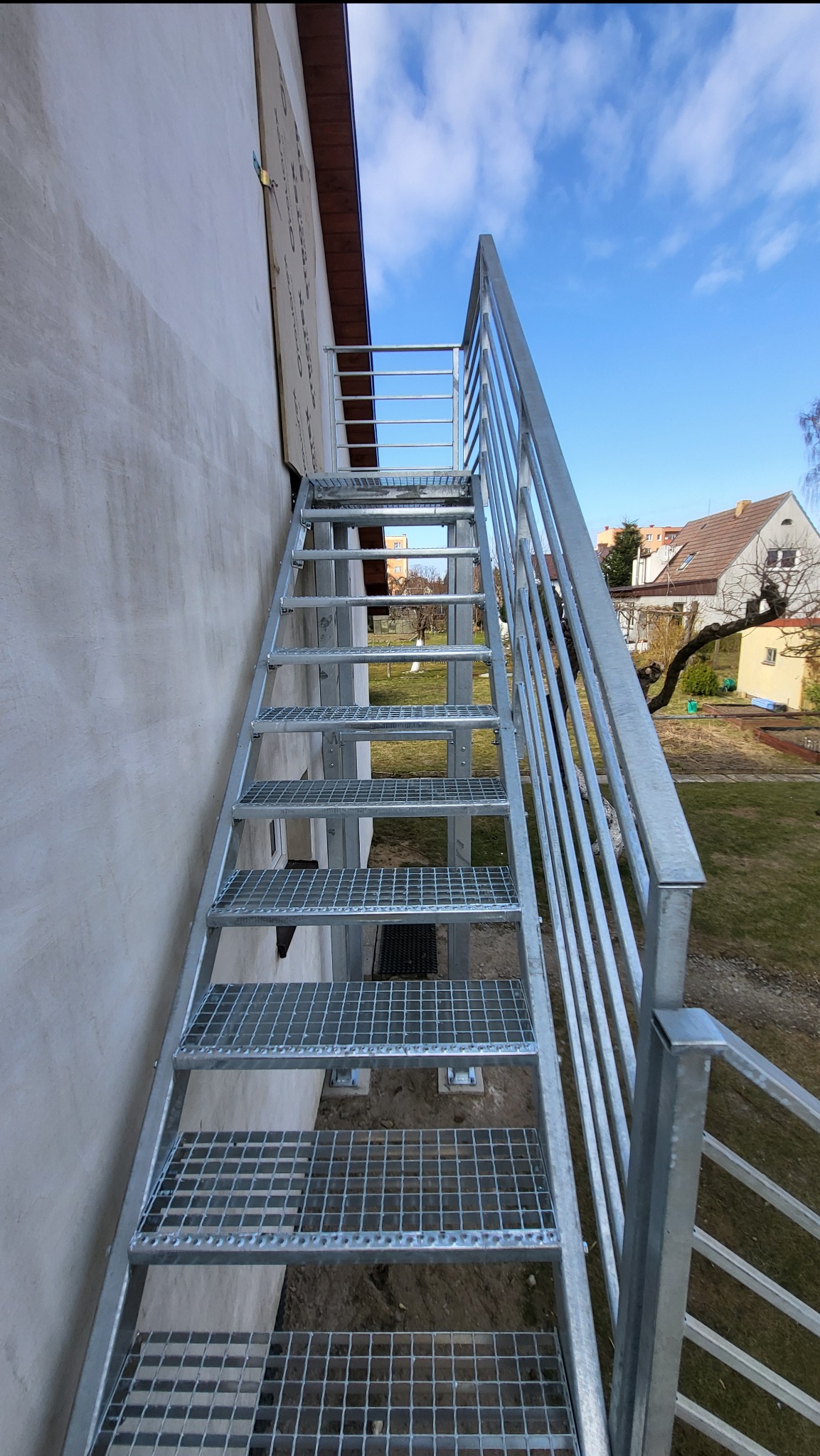 Zewnętrzne schody metalowe z kratowymi stopniami i balustradą, prowadzące wzdłuż ściany budynku. Widok z dołu, błękitne niebo w tle.