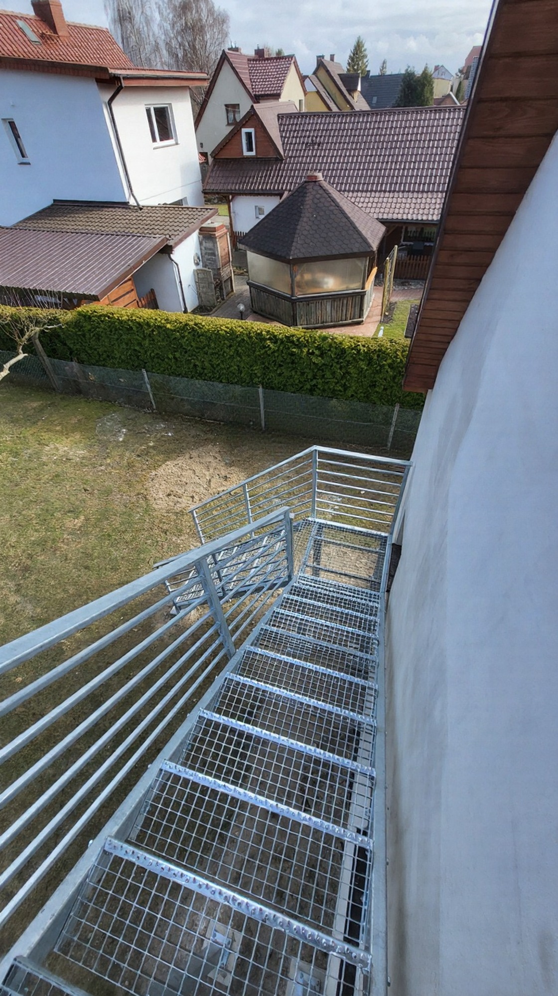 Zewnętrzne schody metalowe z kraty WEMA i balustradą, ocynkowane ogniowo, prowadzące z ogrodu do budynku. Widok z góry, w tle zabudowa jednorodzinna i zieleń.