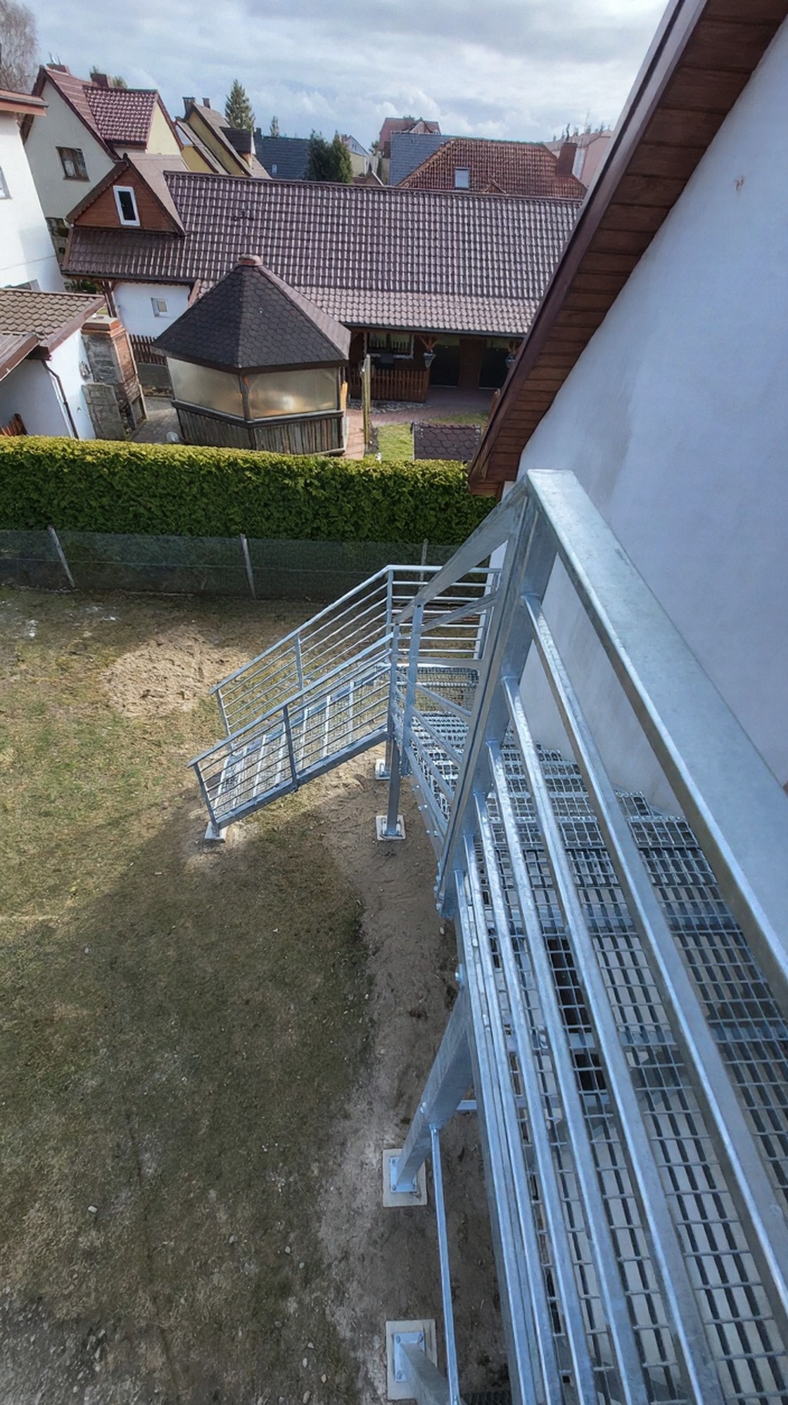 Zewnętrzne schody metalowe z kratowymi stopniami i balustradą, ocynkowane ogniowo, prowadzące z ogrodu do budynku. Widok z góry, na tle zabudowy mieszkalnej.