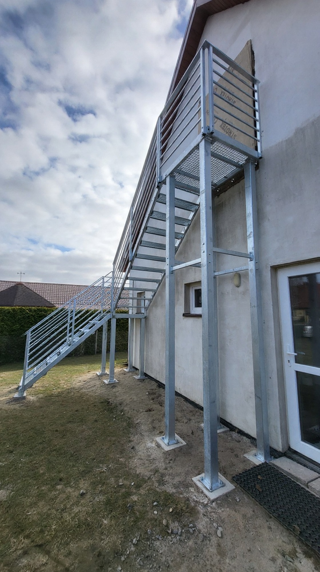 Zewnętrzne schody metalowe z balustradą, ocynkowane ogniowo, prowadzące do wejścia na piętro budynku. Stopnie z kraty antypoślizgowej WEMA.