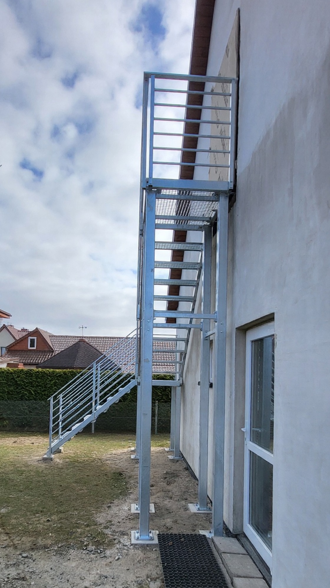 Zewnętrzne schody metalowe z balustradą, ocynkowane ogniowo, prowadzące do drzwi na piętrze budynku. Stopnie z kraty antypoślizgowej WEMA. Widok z boku, pochmurne niebo.