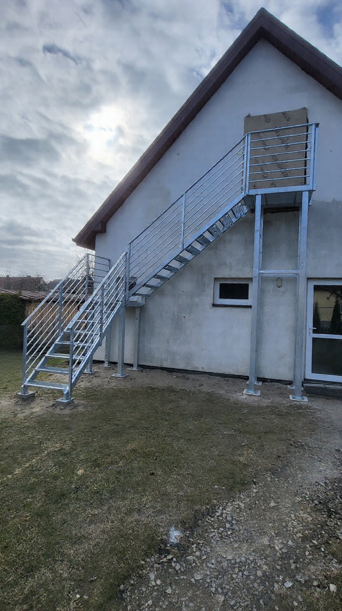 Zewnętrzne schody metalowe z balustradą, ocynkowane ogniowo, prowadzące do drzwi na piętrze budynku. Stopnie z kraty WEMA, słupki i wypełnienie balustrady z profili.