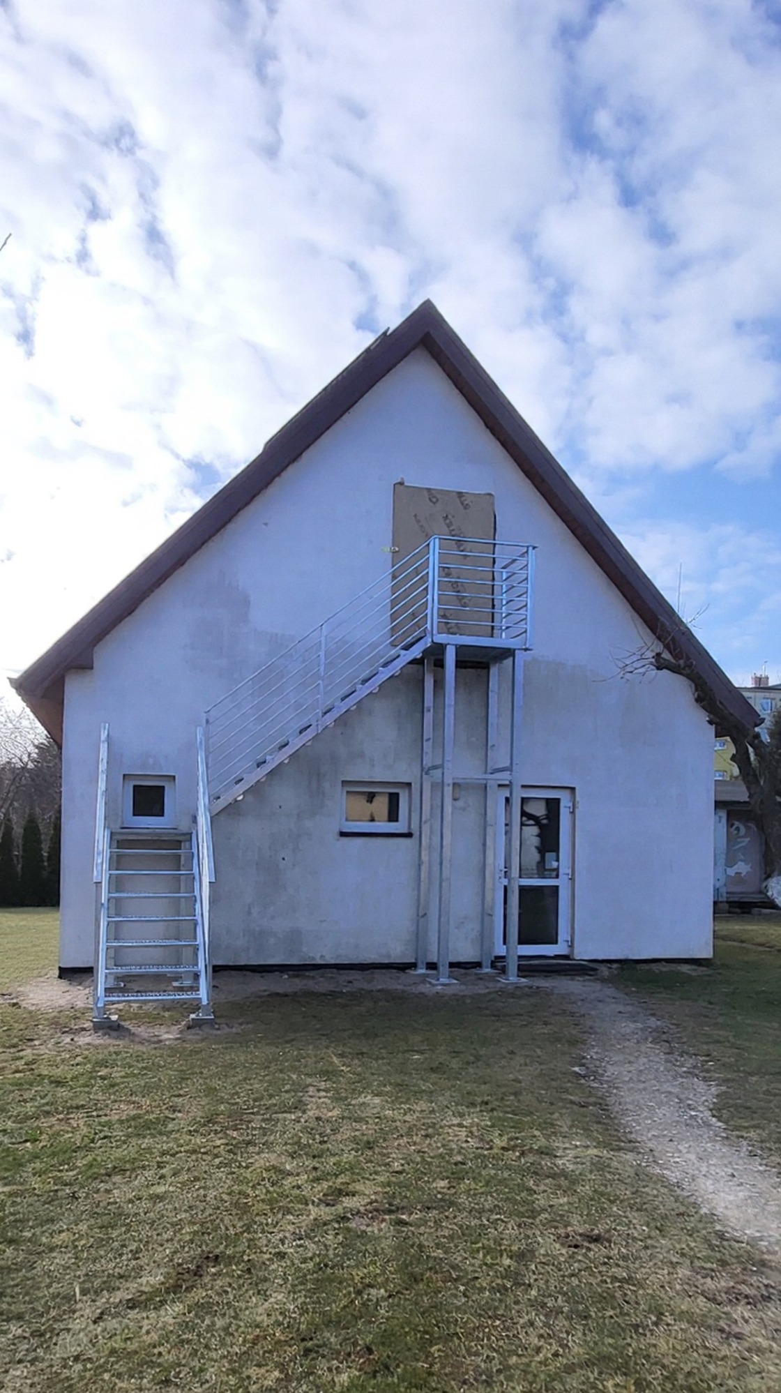 Zewnętrzne schody metalowe z balustradą, prowadzące do drzwi na piętrze budynku. Schody i balustrada w kolorze srebrnym, zamontowane przy białej ścianie budynku.