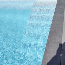 ROYAL POOL PROSTA SPÓŁKA AKCYJNA - Krystalicznie czysta woda w basenie z szarym obramowaniem, widoczny cień osoby po prawej stronie oraz zielone koło do pływania w tle.