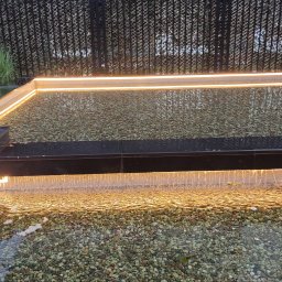 ROYAL POOL PROSTA SPÓŁKA AKCYJNA - Nowoczesny oczko wodne z podświetleniem LED i kaskadą wody spływającą na kamienne dno, otoczone metalową siatką.