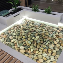 ROYAL POOL PROSTA SPÓŁKA AKCYJNA - Nowoczesny, podświetlany zbiornik wodny z kamieniami, zasilany małą kaskadą, otoczony roślinami w białych donicach i drewnianą kratką.