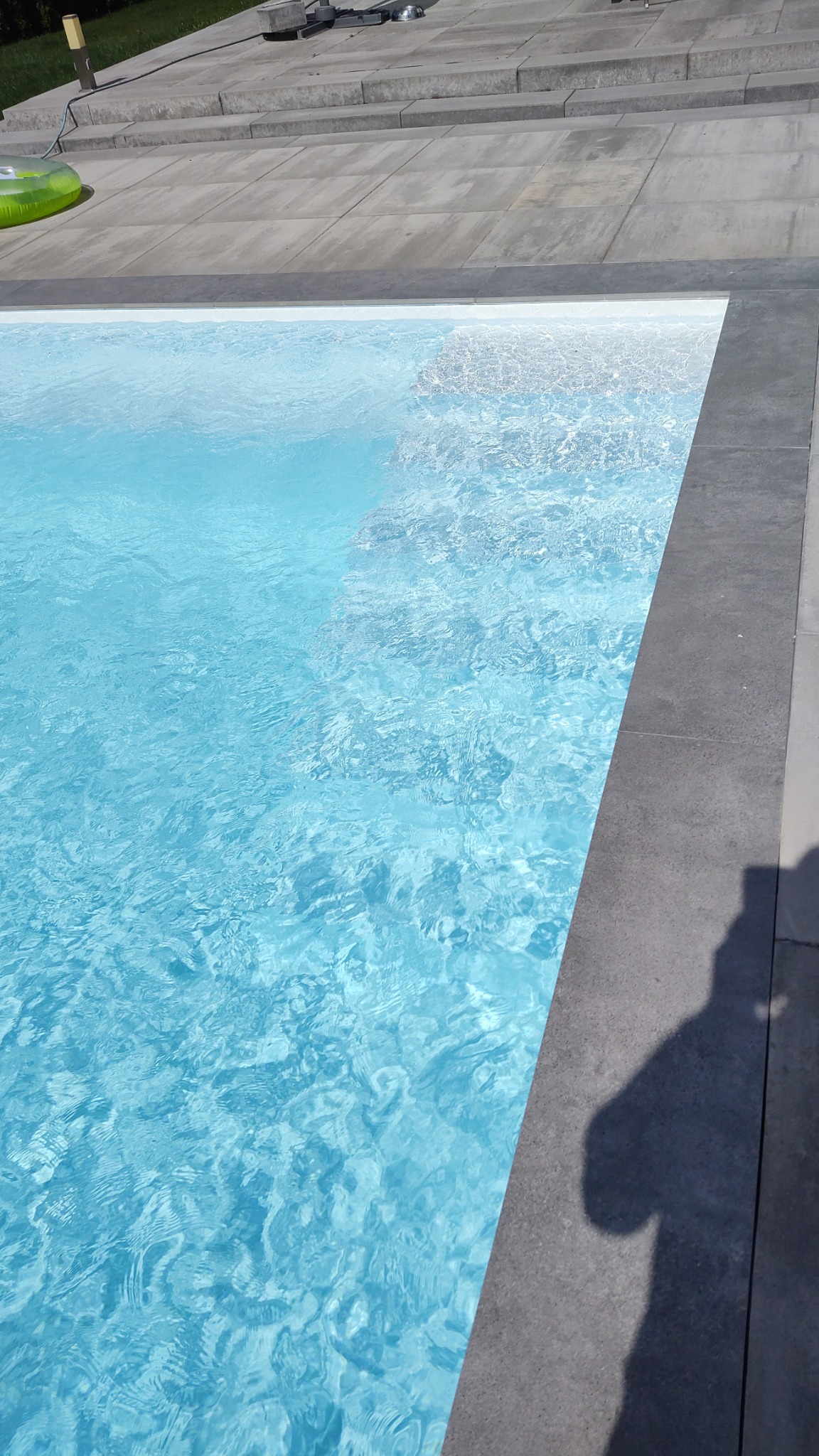 Krystalicznie czysta woda w basenie z szarym obramowaniem, widoczny cień osoby po prawej stronie oraz zielone koło do pływania w tle.