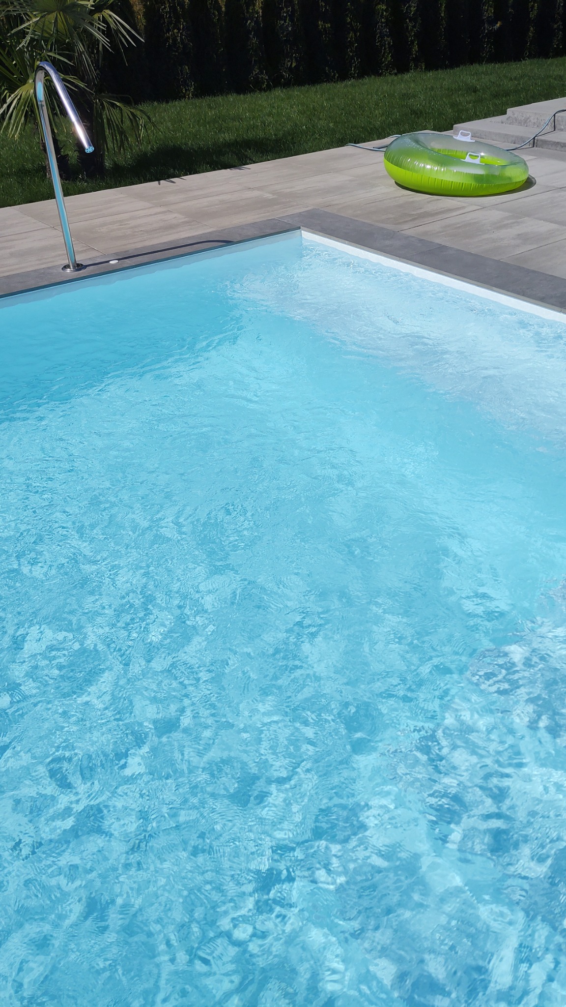 Krystalicznie czysta woda w nowoczesnym basenie z metalową drabinką i zielonym dmuchanym materacem na jasnoszarej, betonowej obudowie basenu, otoczonym zielonym trawnikiem i żywopłotem.