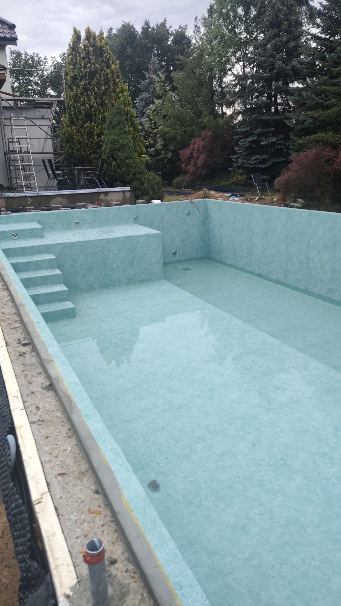 Napełniony wodą basen w trakcie budowy, wyłożony jasnoniebieską folią, z widocznymi schodami i platformą, otoczony zielenią i rusztowaniami.