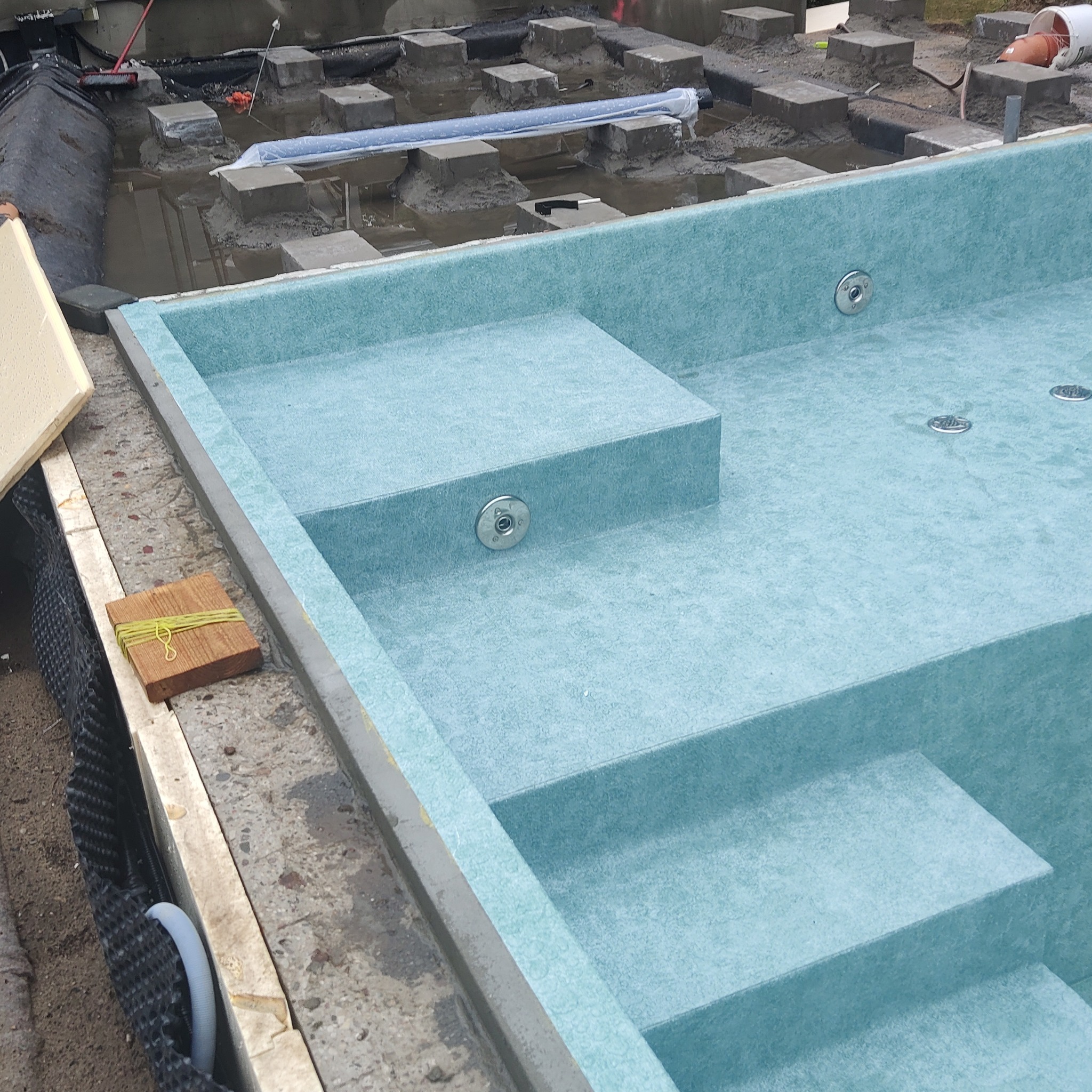 Niebieski basen z włókna szklanego w trakcie instalacji, widoczne schody i dysze, w tle betonowe podpory i elementy konstrukcyjne.