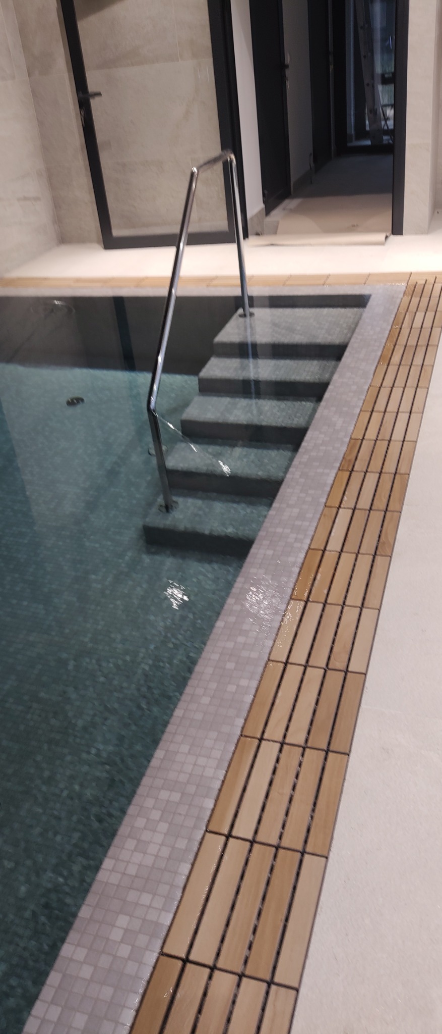 Nowoczesny basen wewnętrzny z metalową poręczą i schodami, wykończony mozaiką i drewnianą podłogą, widok na wejście do budynku.