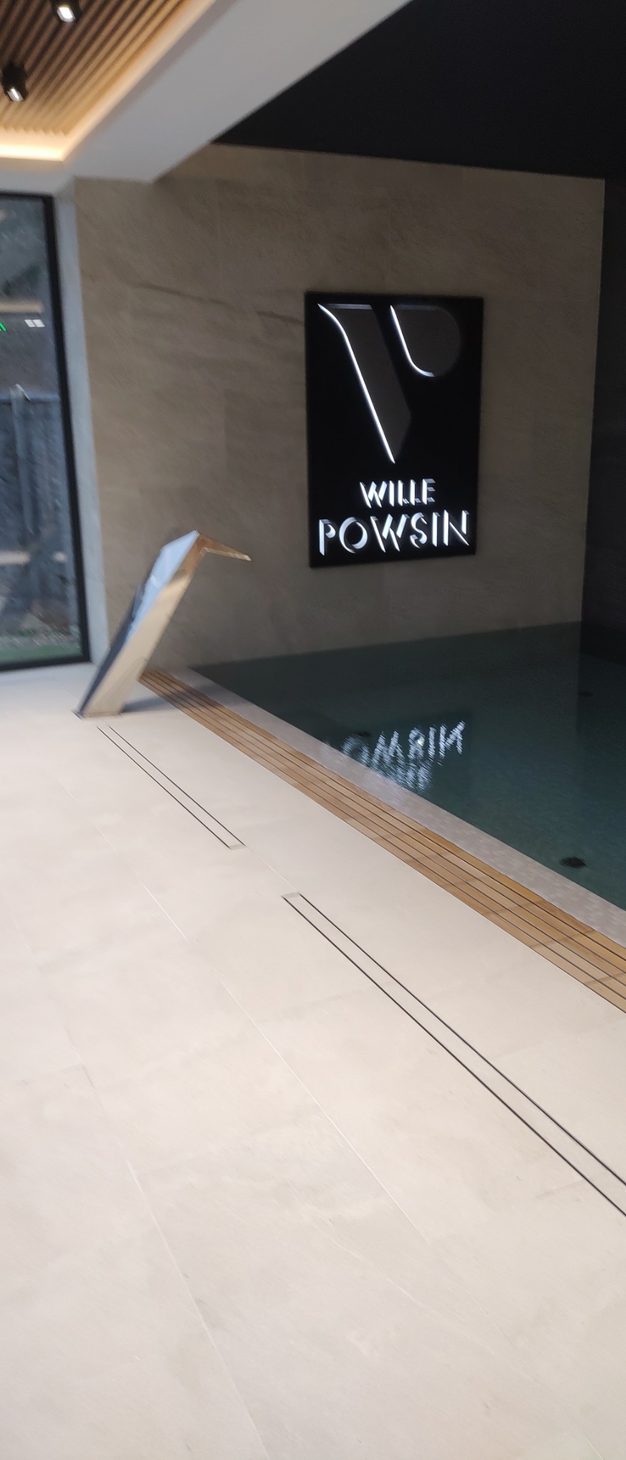 Nowoczesny basen wewnętrzny z minimalistyczną rzeźbą w kształcie ptaka i podświetlanym logo 'Wille Powsin' odbijającym się w wodzie.