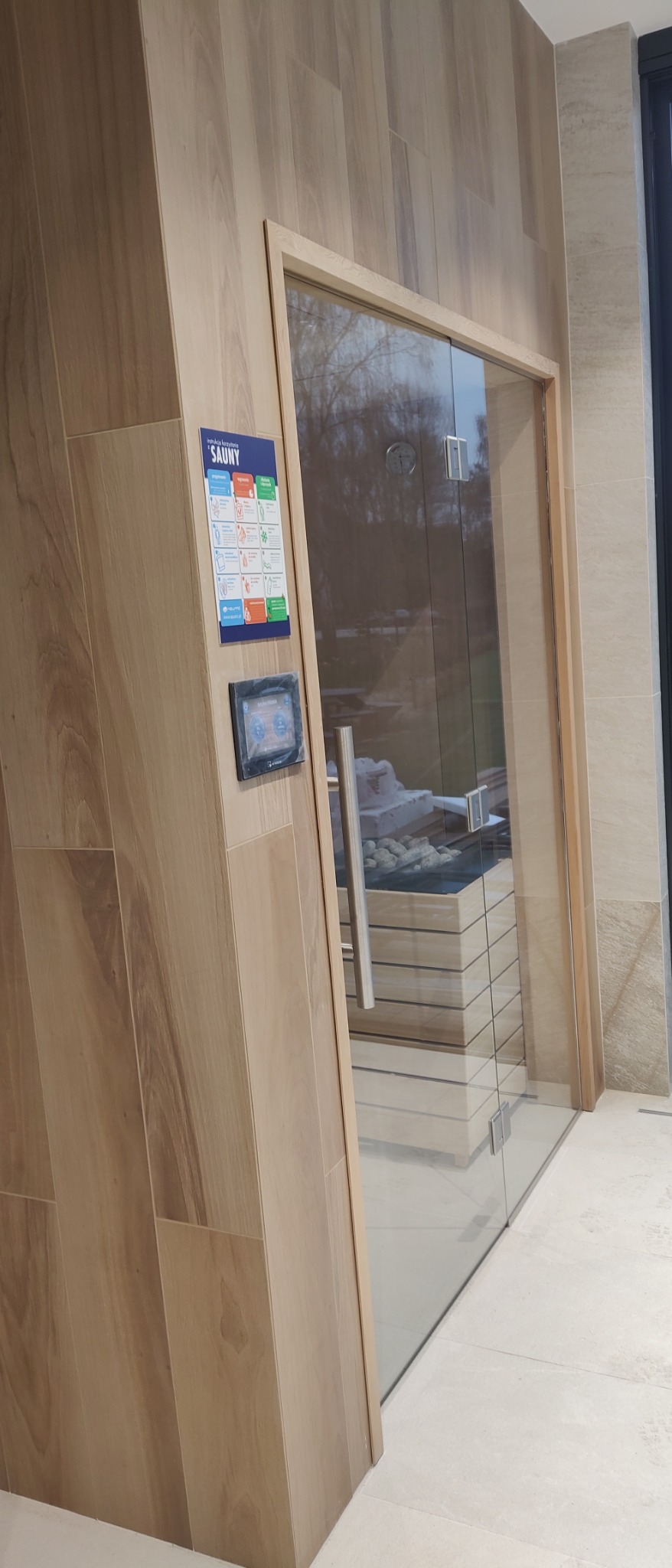 Nowoczesna sauna z drewnianym wykończeniem, szklane drzwi z metalowym uchwytem, panel sterowania i instrukcja obsługi na ścianie, widoczne kamienie wewnątrz sauny.