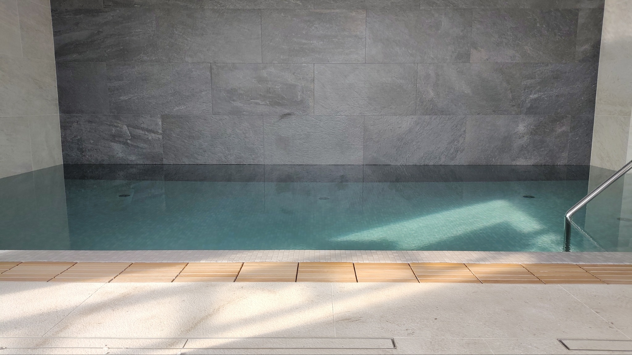 Wnętrze basenu z szarymi płytkami na ścianach, jasnymi płytkami na podłodze i metalową poręczą w rogu.