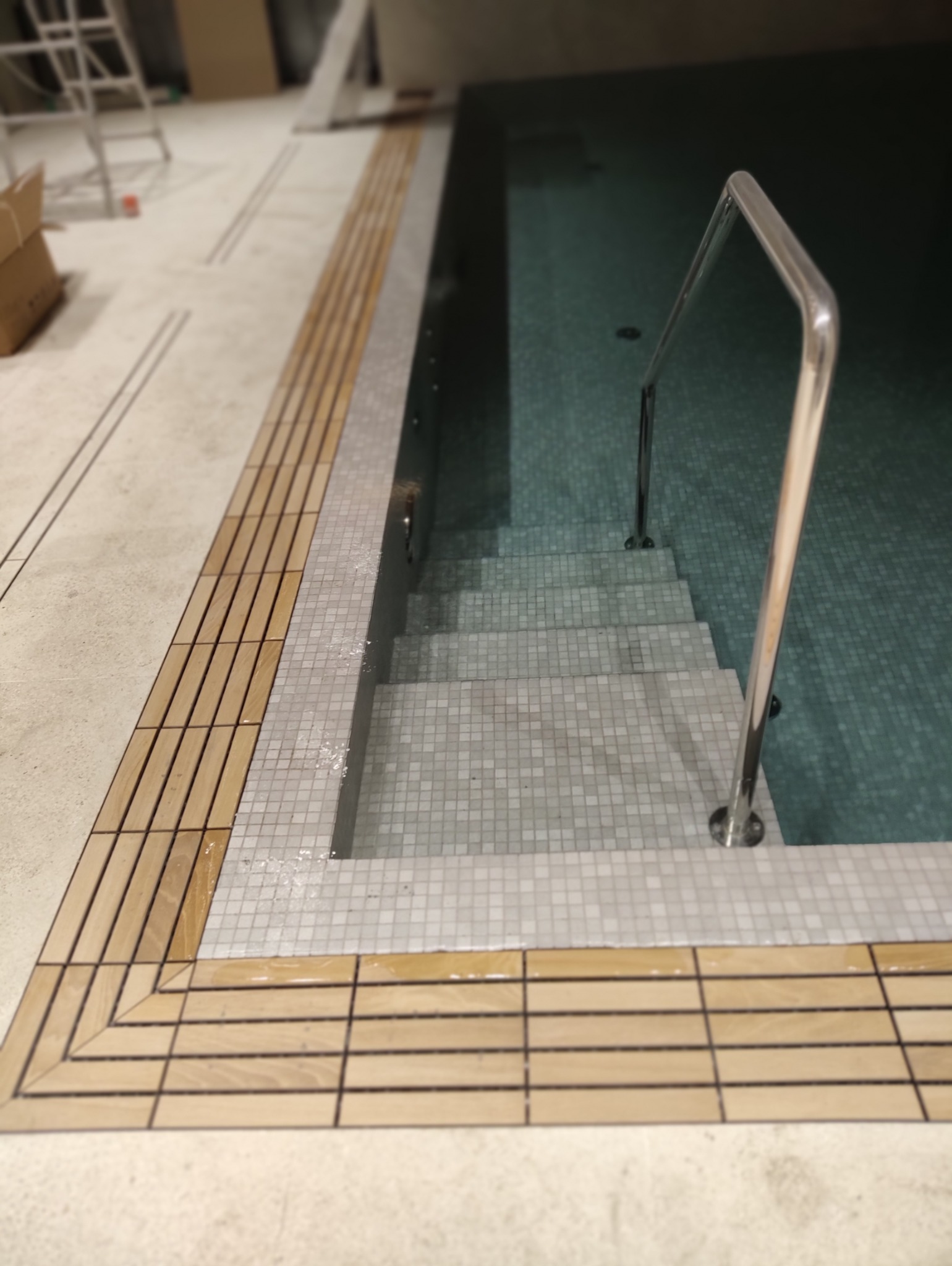 Schody zejścia do basenu wyłożone mozaiką, z metalową poręczą, obrzeże wykończone drewnianymi listwami.