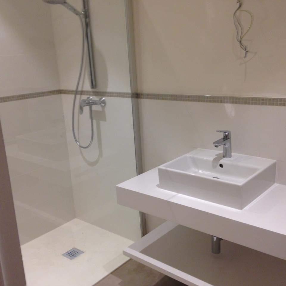 Nowoczesna łazienka z białą kabiną prysznicową typu walk-in, prostokątną umywalką nablatową na białym blacie z półką, wykończona mozaikowym pasem płytek.