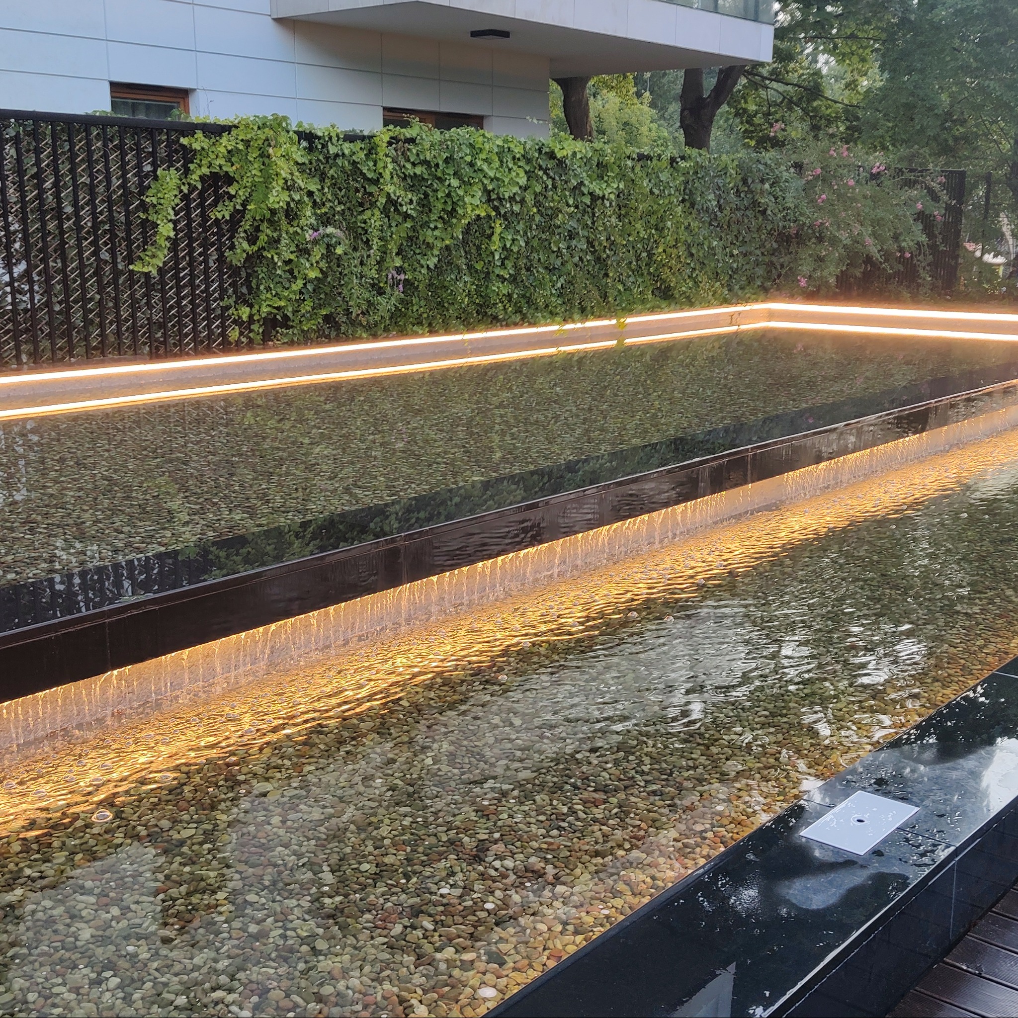 Nowoczesny, podświetlany zbiornik wodny z kaskadą, wypełniony drobnymi kamieniami, otoczony zielenią i czarnym metalowym ogrodzeniem z pnączami.