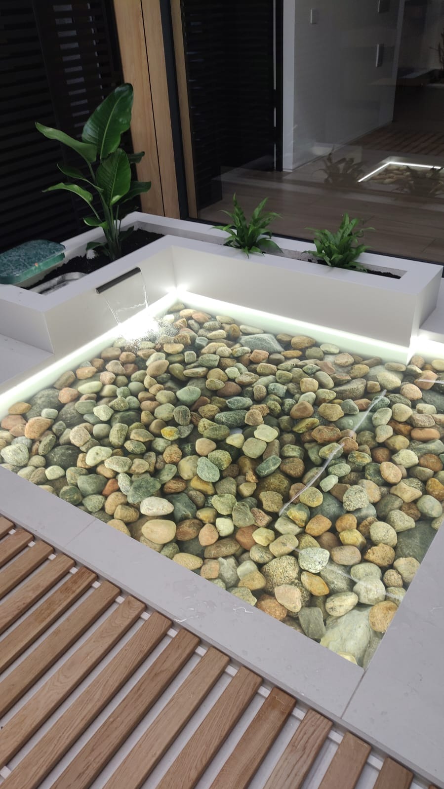 Nowoczesny, podświetlany zbiornik wodny z kamieniami, zasilany małą kaskadą, otoczony roślinami w białych donicach i drewnianą kratką.