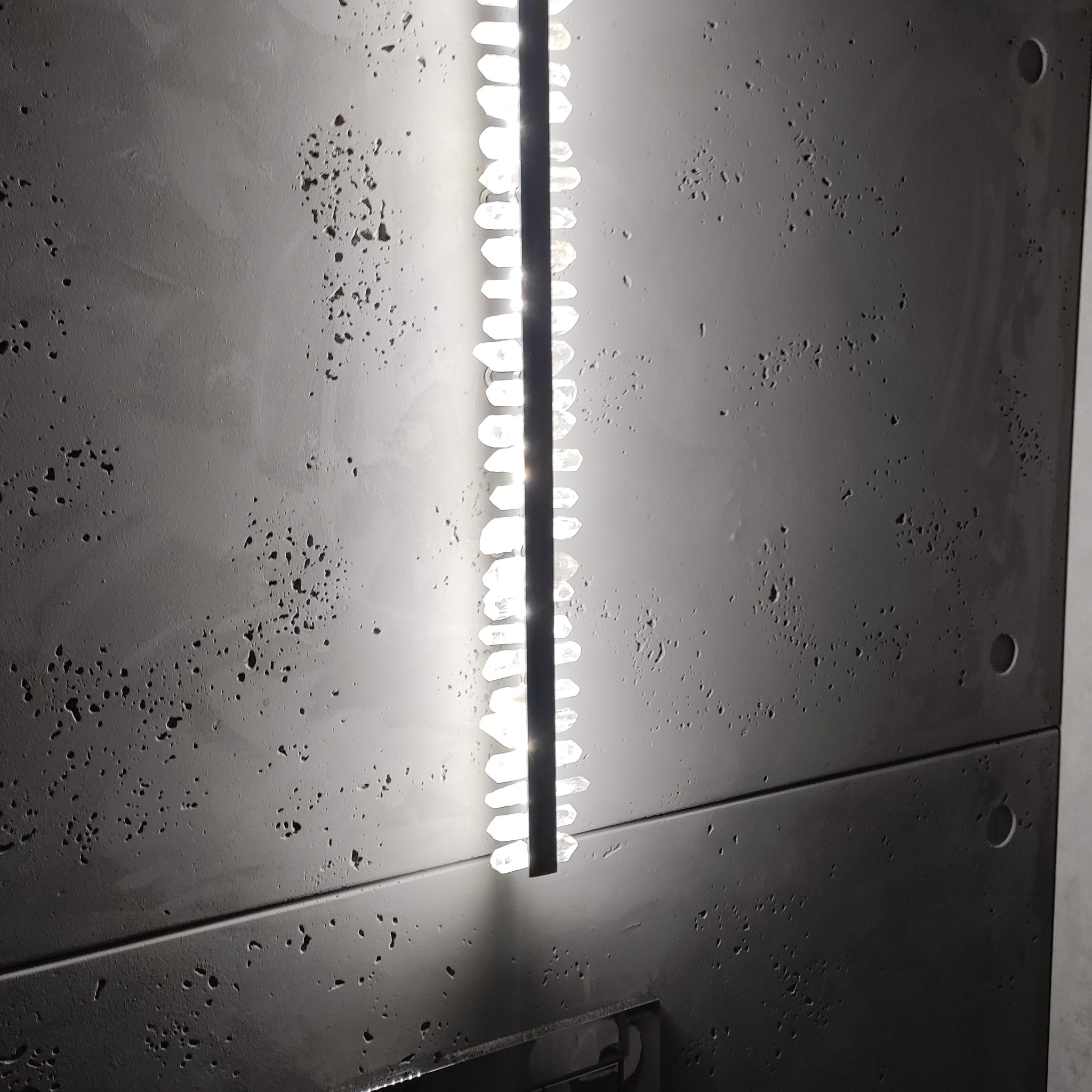 Szara ściana z dekoracyjną, pionową listwą oświetleniową z białymi, geometrycznymi elementami, przypominającymi kryształy.