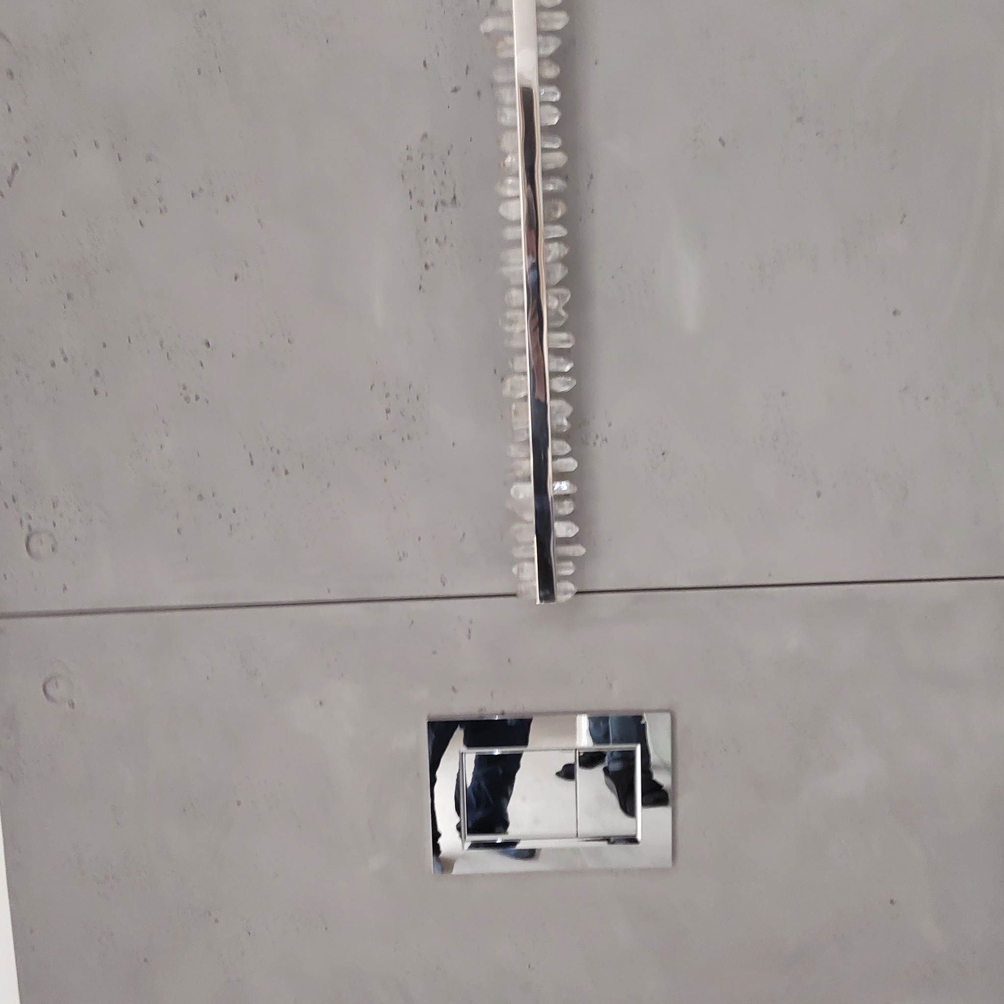 Szara ściana z dekoracyjną, pionową listwą z kryształkami i chromowanym przyciskiem spłuczki WC z odbiciem osoby.