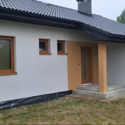 Nowoczesny parterowy dom z szarą dachówką, drewnianymi oknami i drzwiami, oraz jasną elewacją, z widocznym fragmentem fundamentu zabezpieczonego folią.