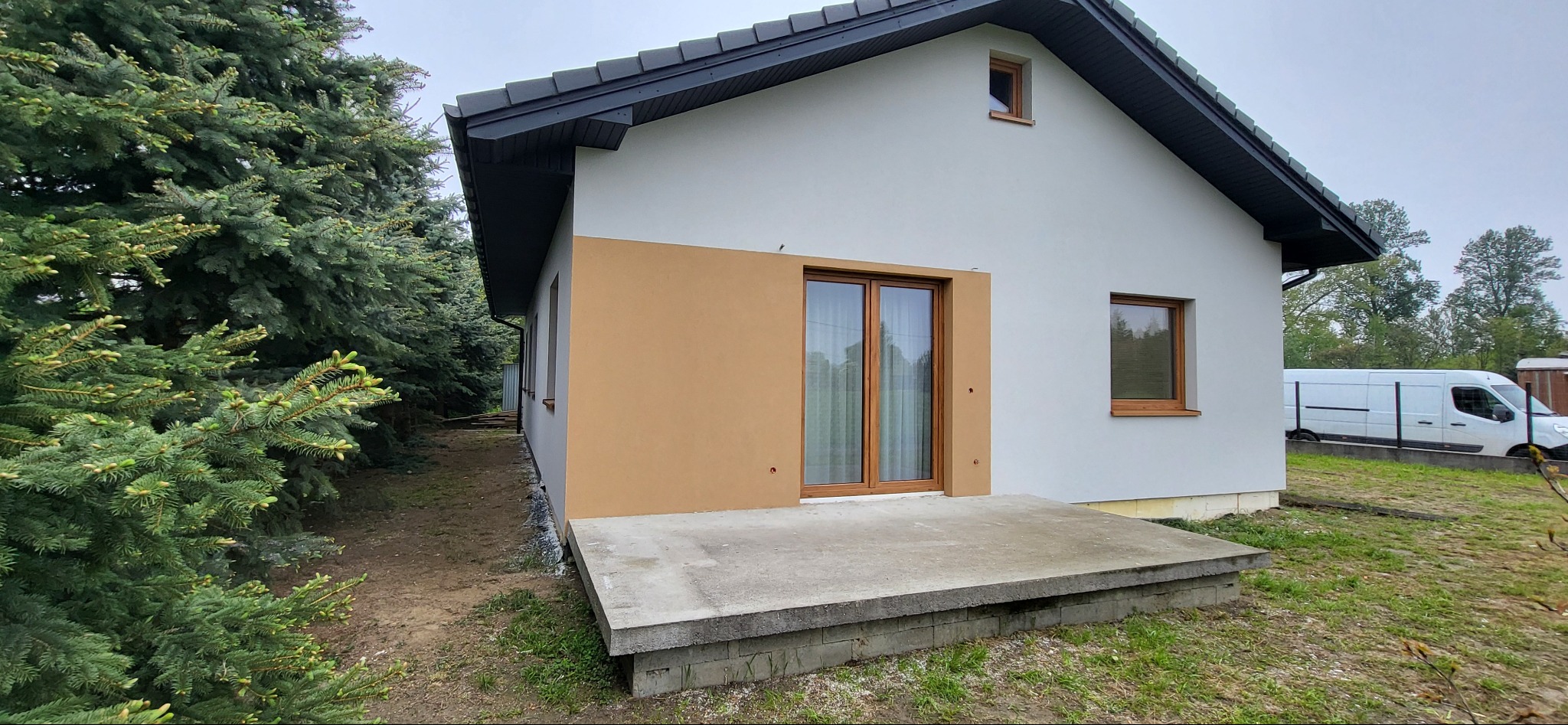 Nowoczesny dom jednorodzinny z płaskim dachem i elewacją w dwóch odcieniach, widoczny taras betonowy i okna z drewnianą ramą, na tle zielonych drzew i białego samochodu dostawczego.