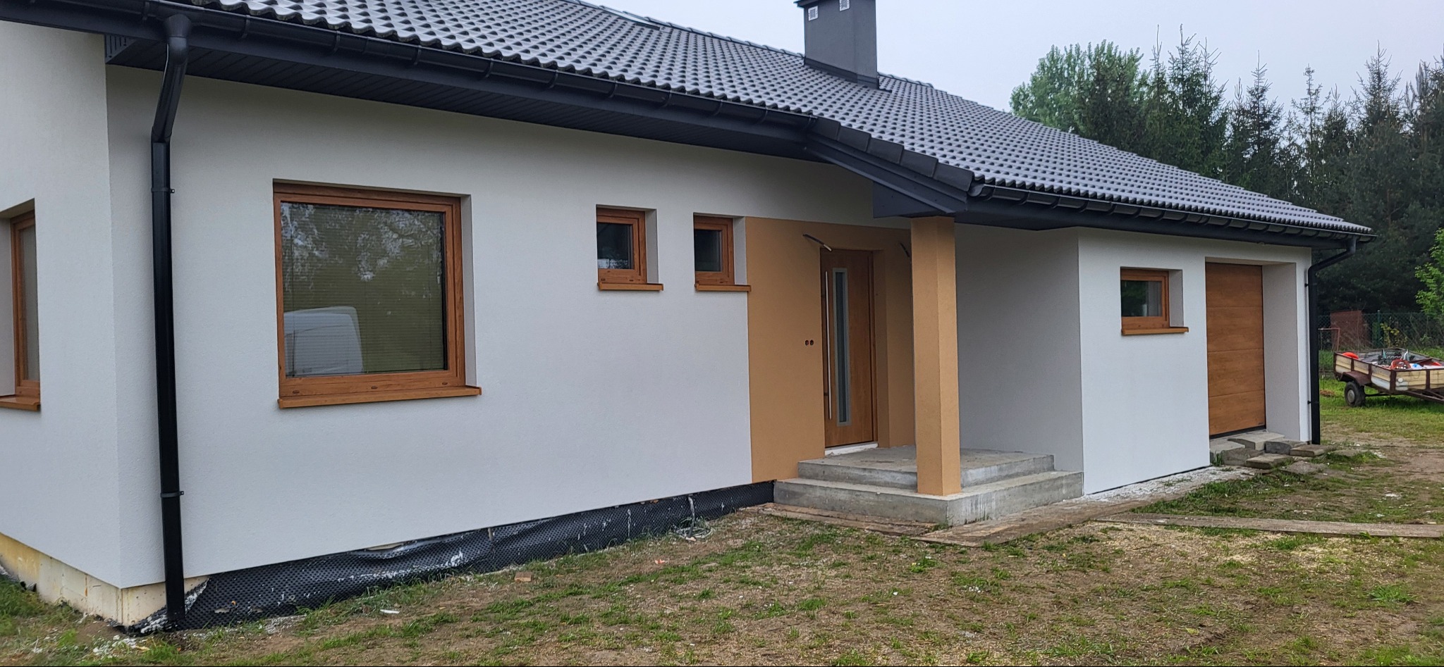 Nowoczesny parterowy dom z szarą dachówką, drewnianymi oknami i drzwiami, oraz jasną elewacją, z widocznym fragmentem fundamentu zabezpieczonego folią.