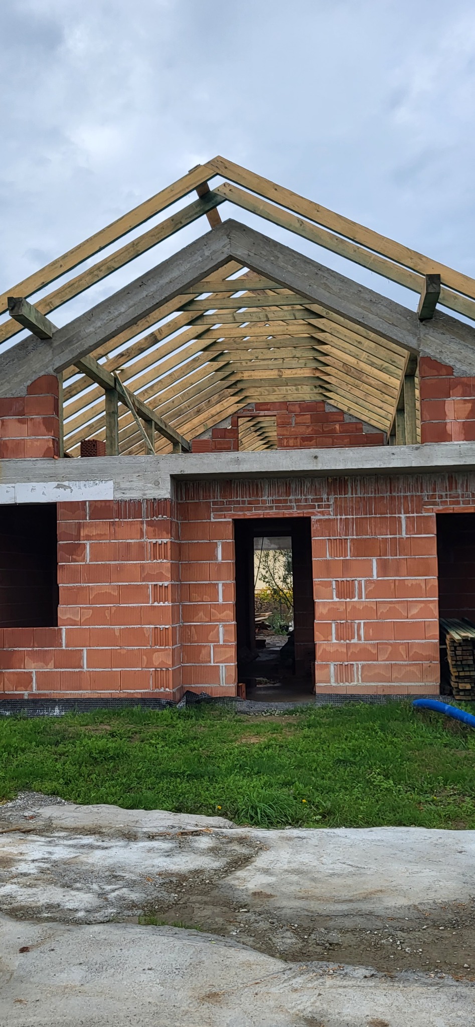 Surowy stan budynku z czerwonej cegły z widoczną drewnianą konstrukcją dachu i betonowym wieńcem, widok na podwórko przez otwór drzwiowy.