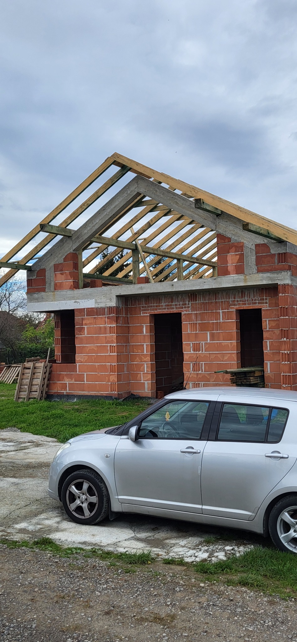 Surowy stan domu z czerwonej cegły z drewnianą konstrukcją dachu, widok z zewnątrz, na pierwszym planie zaparkowany srebrny samochód osobowy.