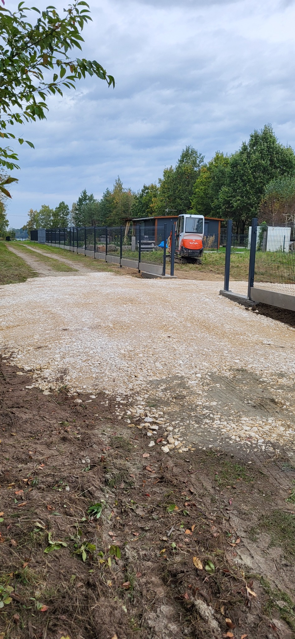 Nowo wyłożona, jasna, kamienista nawierzchnia drogi dojazdowej, z widocznym fragmentem ogrodzenia z betonową podmurówką i siatką, w tle zaparkowana mała koparka.