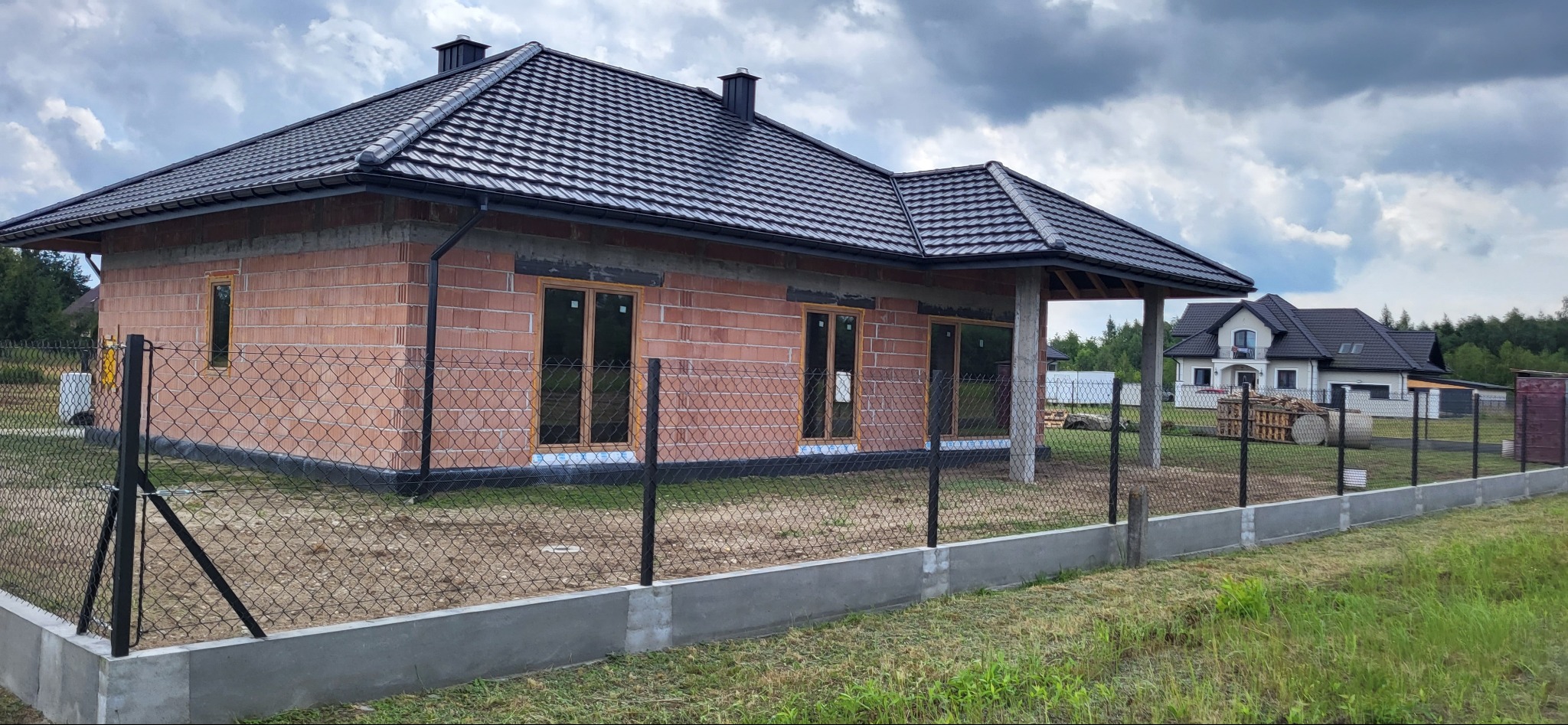 Niedokończony dom jednorodzinny z cegły z czarnym dachem, widoczne okna bez parapetów, ogrodzenie z siatki, w tle drugi dom w budowie.