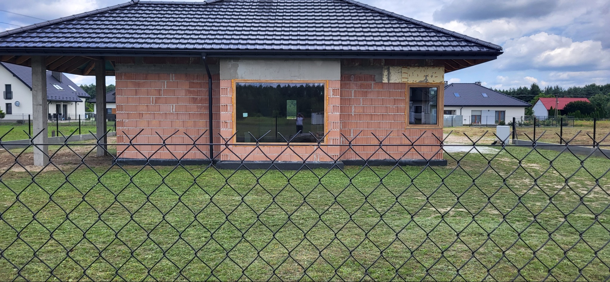 Budynek w trakcie wznoszenia z czerwonej cegły, widoczne okna wstawione w surowy mur, w tle inne domy i zieleń, całość ujęta przez siatkę ogrodzeniową.