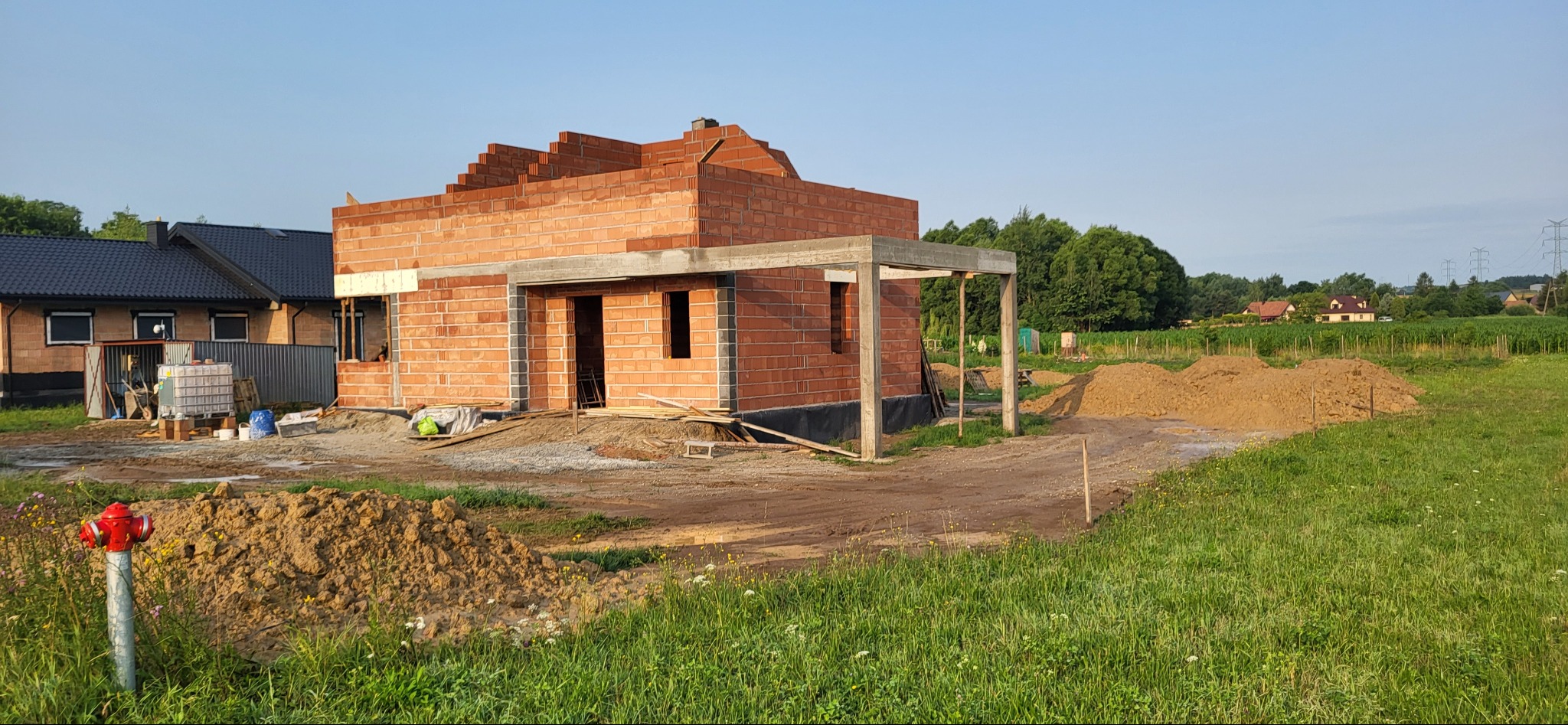 Wznoszony budynek z czerwonej cegły z betonowym zadaszeniem tarasu w surowym stanie, widok na plac budowy, trawa i hydrant w pierwszym planie.