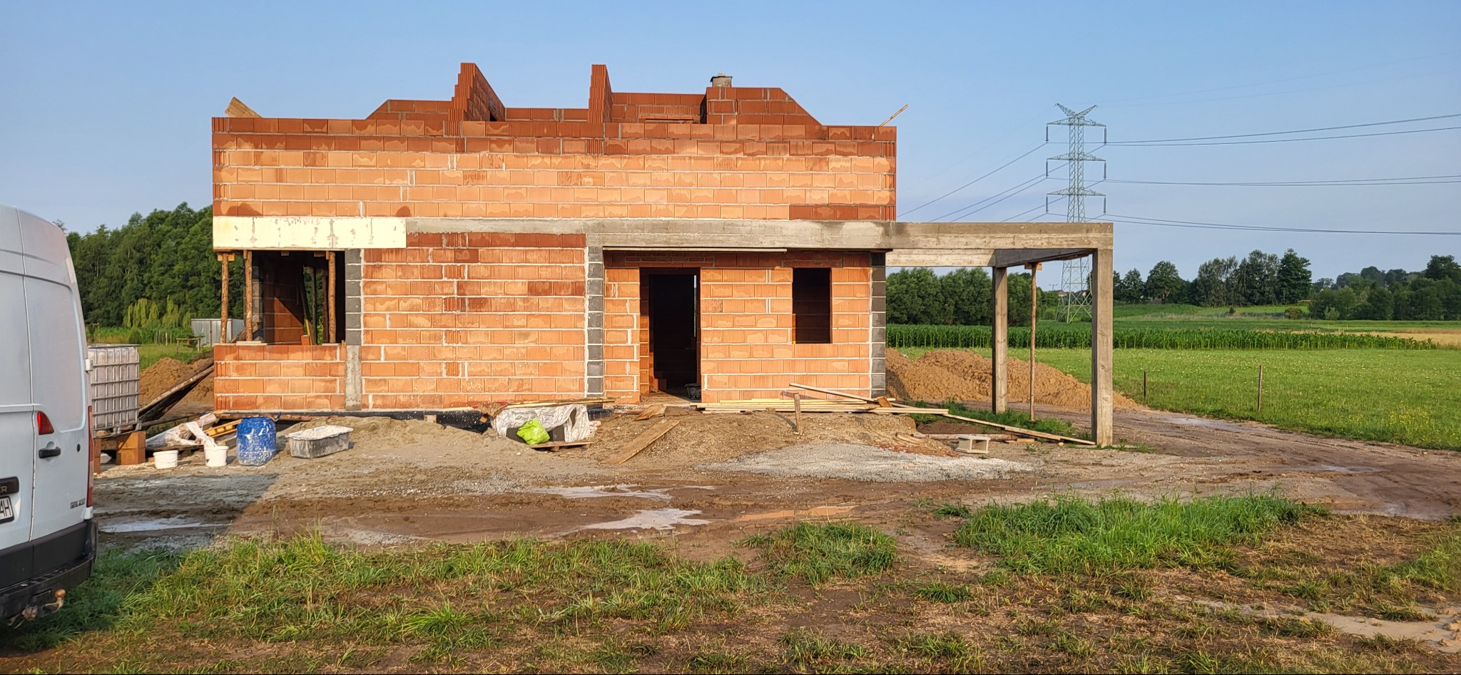 Surowy stan budynku z czerwonej cegły, widoczne otwory okienne i drzwiowe, z elementami szalunku i stertą piasku w tle, częściowo widoczny biały samochód dostawczy po lewej stronie kadru.