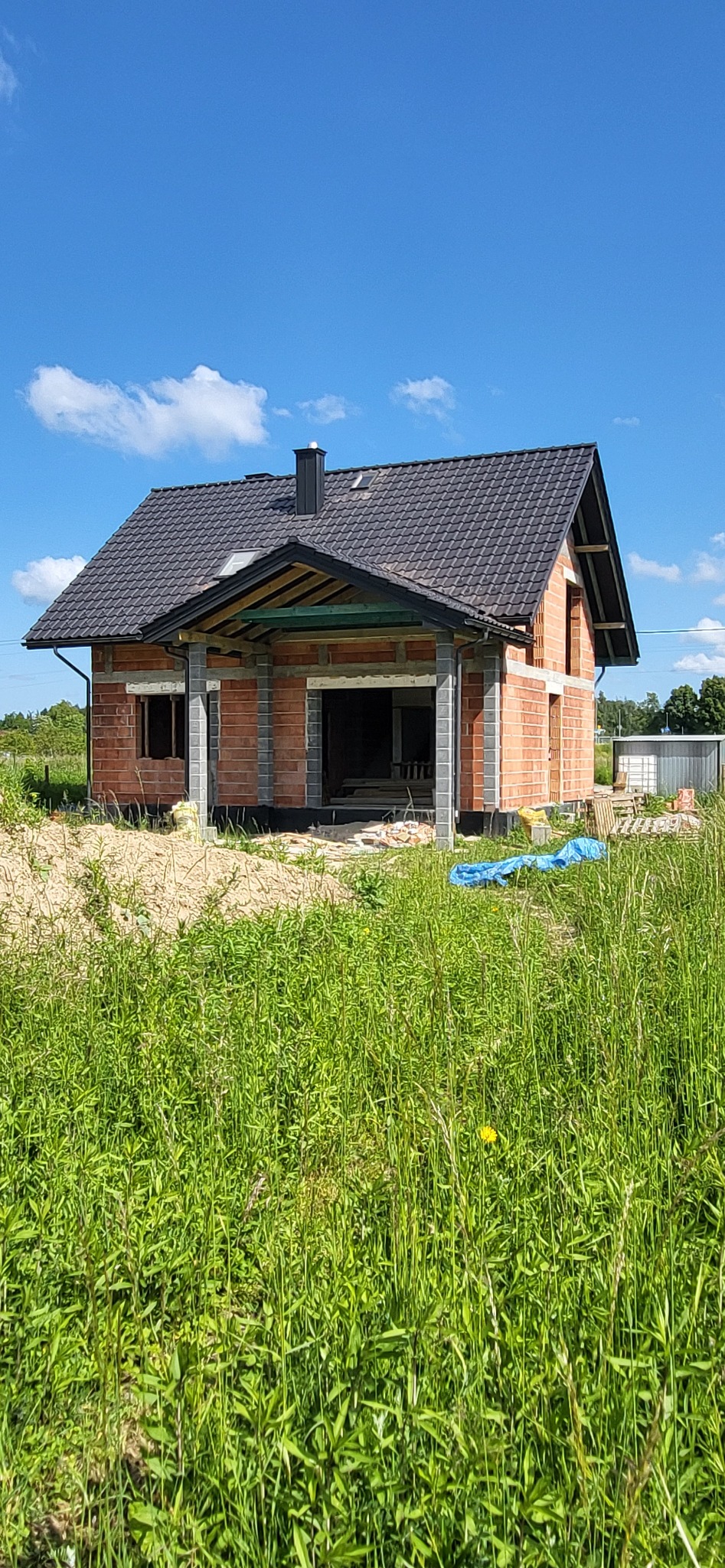 Surowy stan domu jednorodzinnego z cegły i pustaków, z czarnym dachem z dachówki, widoczny komin i podpora werandy, otoczonego wysoką trawą i piaskiem.