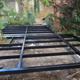 Metalowa konstrukcja pergoli w trakcie montażu nad strumieniem, widoczne czarne profile i fragment zieleni