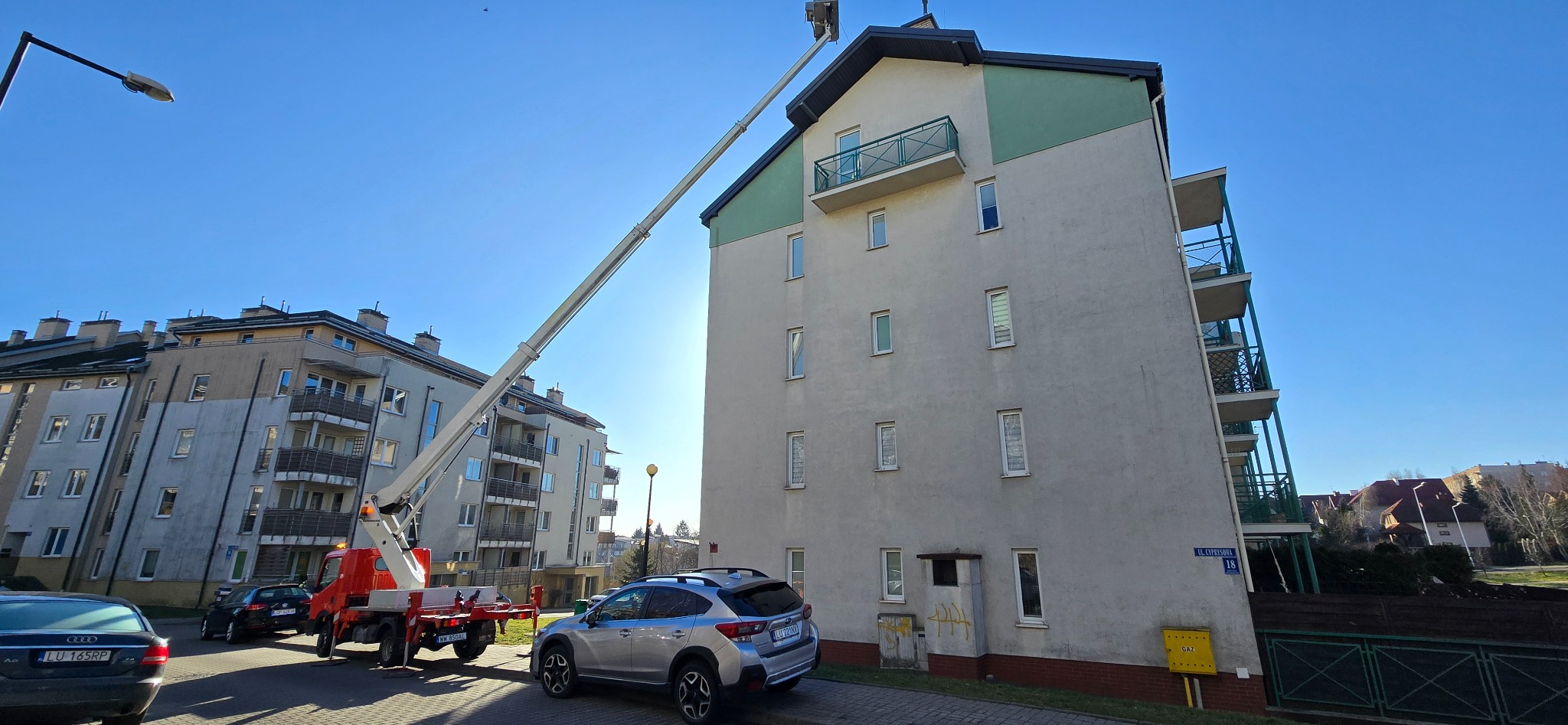 Podnośnik koszowy na tle bloku mieszkalnego z zielonymi elementami, z zaparkowanym srebrnym samochodem osobowym na pierwszym planie i błękitnym niebem w tle.