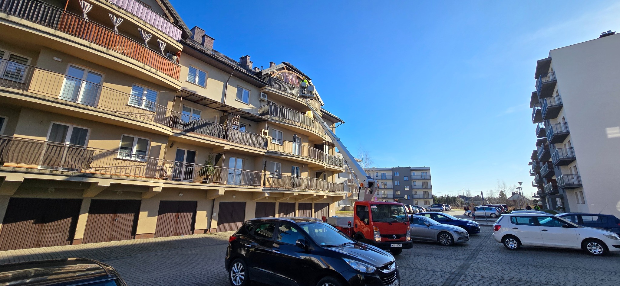 Pracownik na podnośniku koszowym przy dachu budynku wielorodzinnego z balkonami, prawdopodobnie wykonuje prace dekarskie.