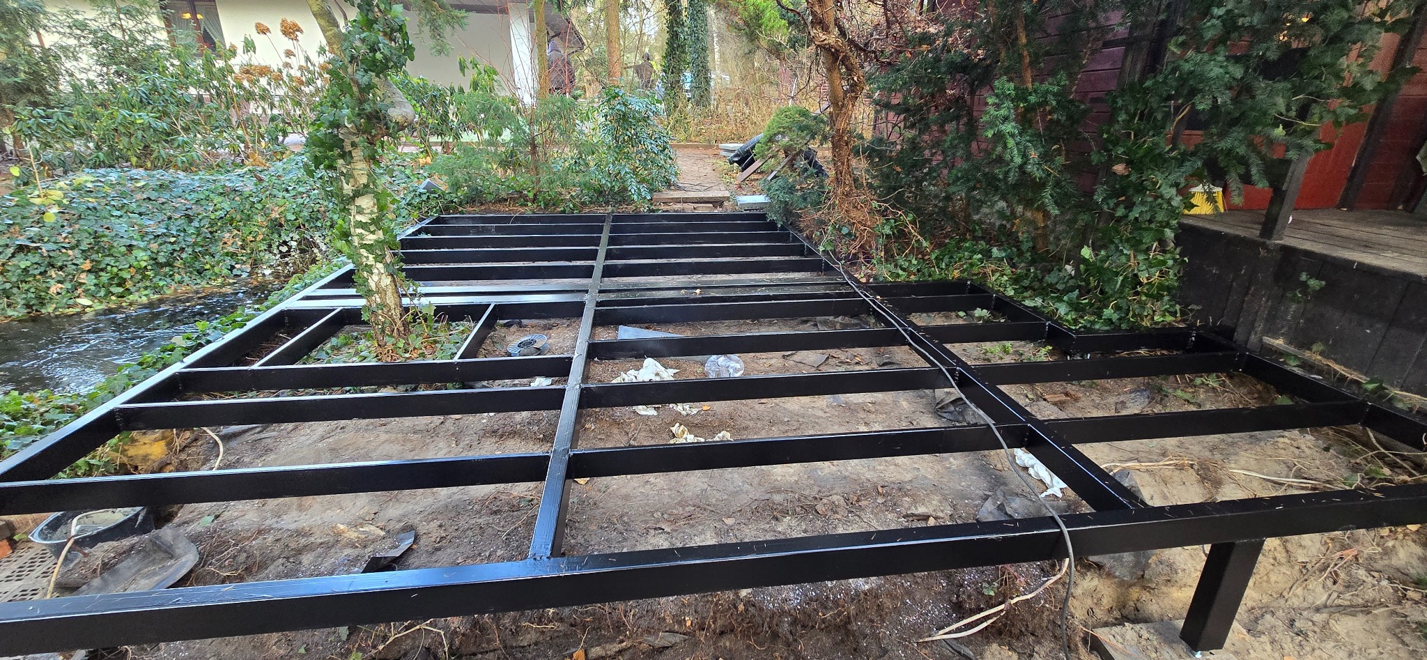 Metalowa konstrukcja pergoli w trakcie montażu nad strumieniem, widoczne czarne profile i fragment zieleni