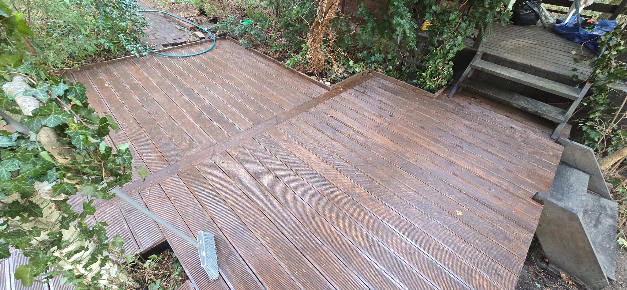 Brązowy taras z desek kompozytowych, częściowo mokry, z widocznymi schodami i szczotką do zamiatania, otoczony zielenią.